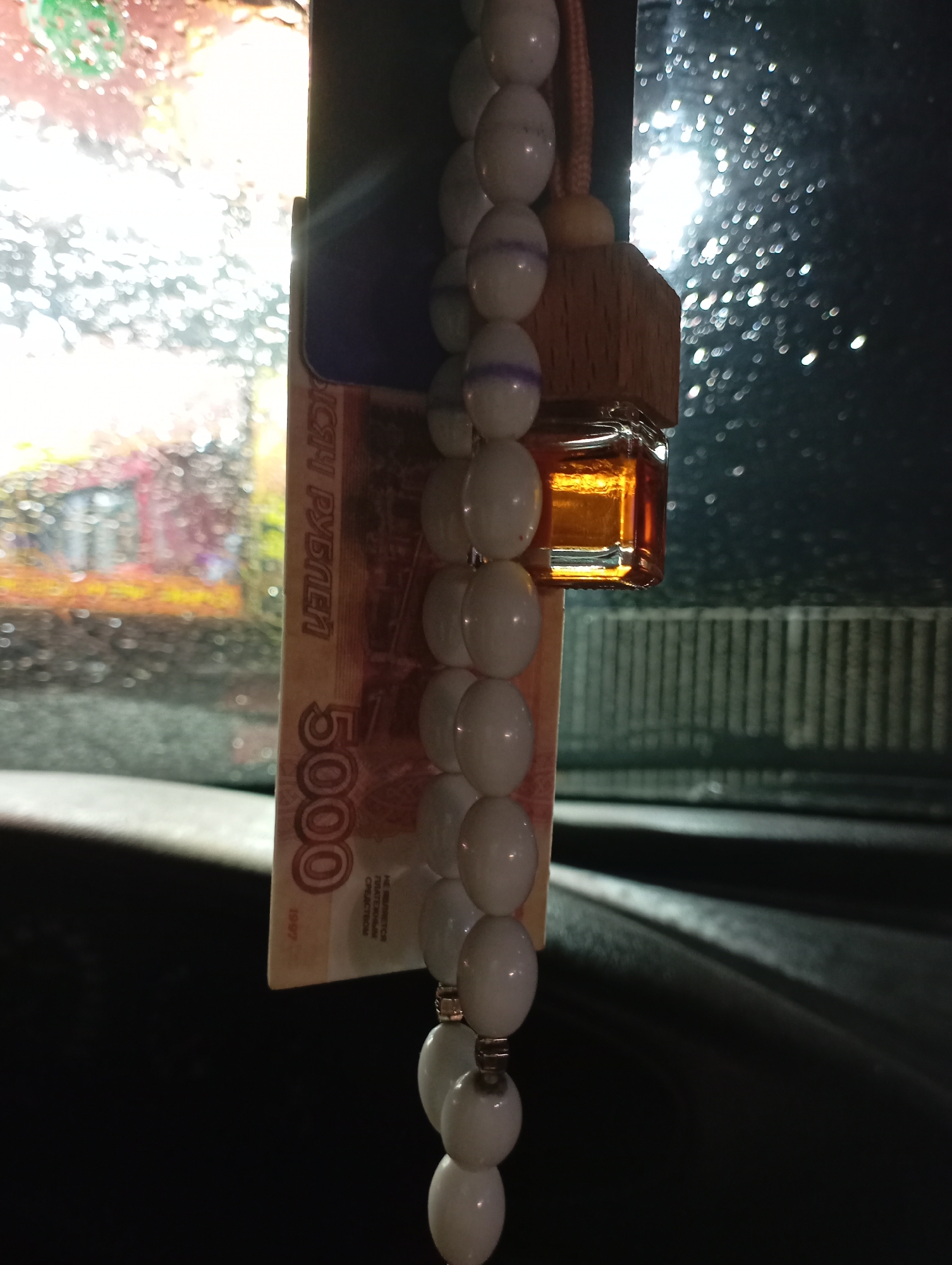 The image shows a 5000 Russian Ruble banknote hanging from a car rearview mirror. The banknote is behind an air freshener and a string of white beads. Through the rain-streaked windshield, the blurred lights of the Industrialny city are visible.