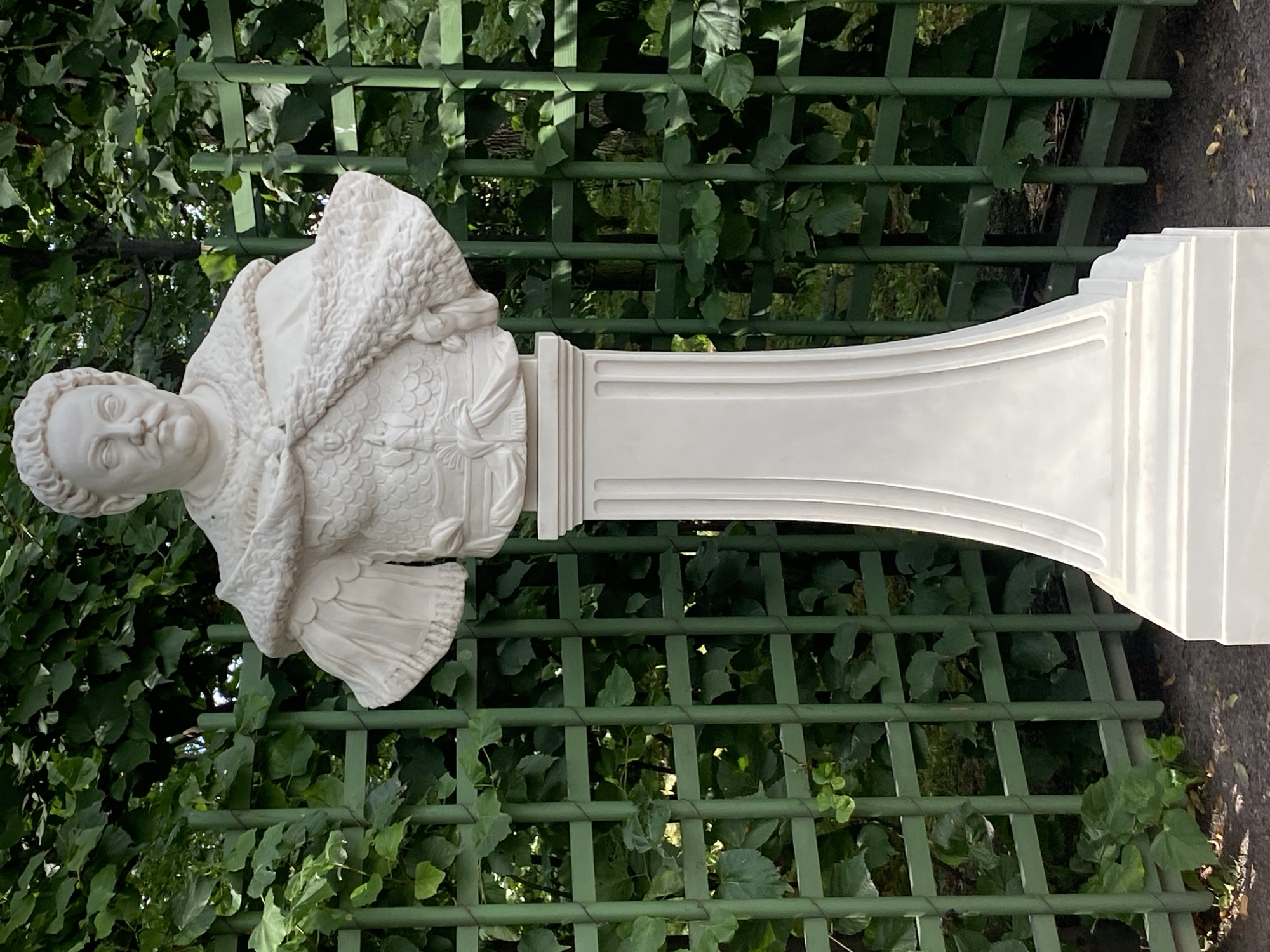 A white marble bust of a man with a mustache, wearing ornate armor and a fur-lined cloak, stands on a fluted white pedestal. The bust is positioned in front of a green lattice fence covered in ivy. The background consists of dense green foliage and trees. The lighting suggests a daytime scene, possibly outdoors in a park or garden. This statue is located in the Summer Garden in Saint Petersburg, Russia, specifically in front of the Hermitage Museum.