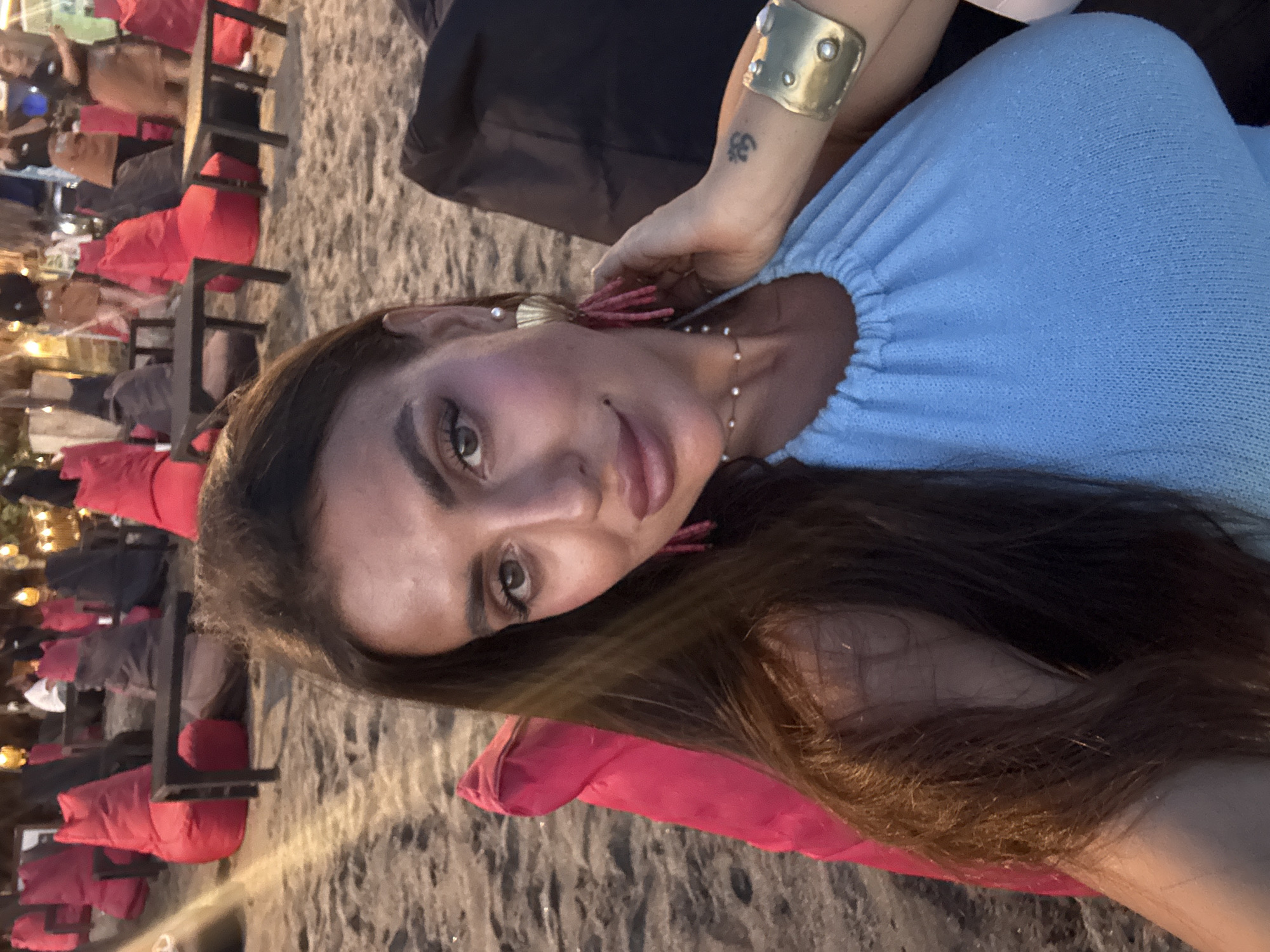 The image shows a woman at sunset on a beach in North Kuta, Indonesia. She is sitting on a pink beanbag chair and wearing a light blue shirt, coral earrings, and a pearl necklace. She is also wearing a gold bracelet on her upper arm. In the background, there are other people sitting on beanbag chairs at tables.