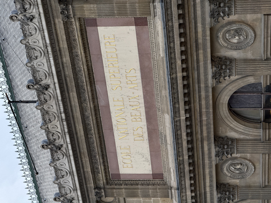 This close-up image displays the upper facade of the École Nationale Supérieure des Beaux-Arts, situated in Paris, France. The architectural details are classical, primarily executed in grey stone with accents of reddish-brown marble. At the top, a grey slate mansard roof is visible, adorned with an ornate dark metal cresting and a slender vertical mast. Below the roofline, an elaborate stone frieze features scrolled patterns and integrated sculptural heads. The central element is a large rectangular plaque, framed by stone molding, with a light-colored stone field bordered in reddish-brown marble. Embossed in gold-colored, capitalized letters on this plaque is the text "ECOLE NATIONALE SUPÉRIEURE DES BEAUX-ARTS". Beneath this, the facade continues with further ornamented friezes, fluted pilasters capped with Corinthian capitals, and circular stone medallions featuring bas-relief profiles. A segment of an arched window with dark panes is visible at the bottom. No individuals are present. The ambient light suggests an overcast sky.