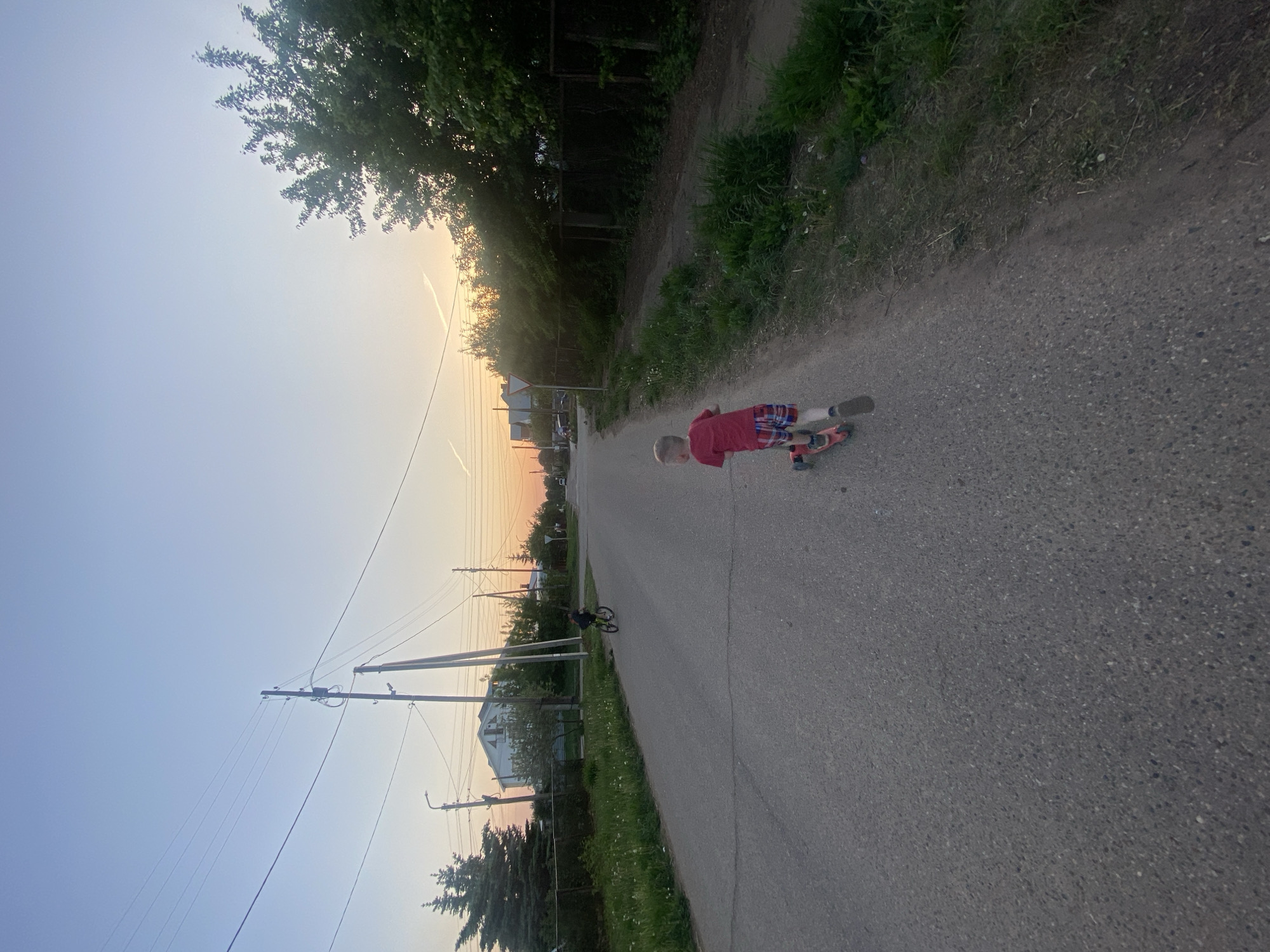 A child in a red shirt and plaid shorts is riding a scooter on a paved road. The scene is located in Прикамский, Russia, as evidenced by the presence of infrastructure and architecture typical of the region. Power lines run parallel to the road on the left side. Trees and greenery line both sides of the road. The sky transitions from blue to orange near the horizon, suggesting either sunrise or sunset. A second individual, a cyclist, is visible further down the road.