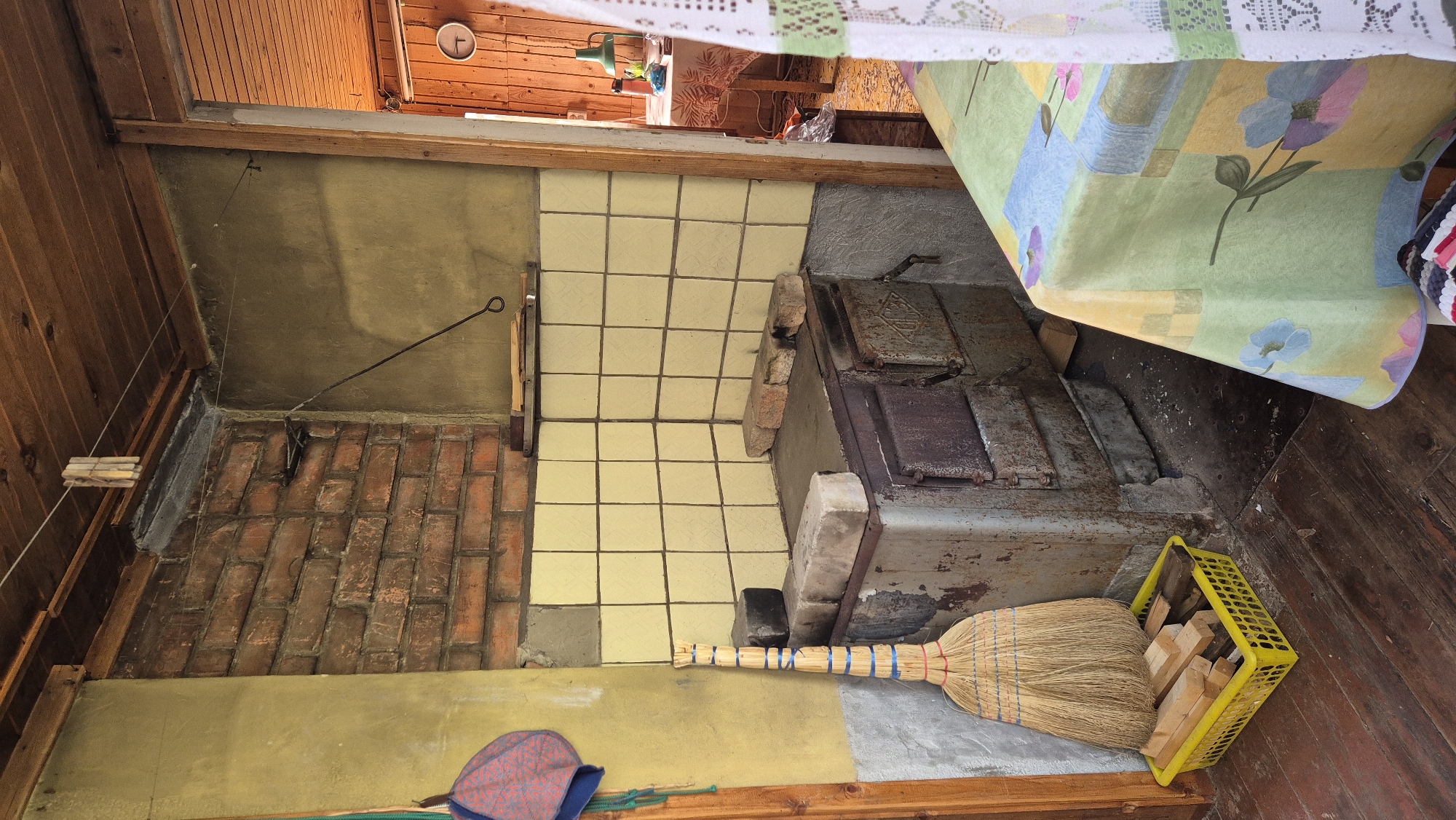 The image depicts a rustic interior scene, likely in a rural or older dwelling, possibly in the village of Mashezero, Russia. The foreground features a worn wooden floor. To the left, a broom made of straw stands upright in a bright yellow plastic basket filled with cut firewood. Next to the broom, a portion of a red and blue patterned hat hangs from a wooden doorframe.
Dominating the center of the image is an old, metal stove with a tiled backsplash. The stove appears to be made of cast iron with visible doors and a flat top. Several bricks are placed on top of the stove. The tiled area behind the stove is a pale yellow color. Above the tiled area, a brick wall is visible, with a thin black metal rod extending from the wall towards the right.
To the right of the stove, a table is partially visible, covered with a tablecloth featuring a colorful floral pattern in shades of green, yellow, blue, and pink. A white lace curtain hangs on the right edge of the image. Beyond this, an open doorway reveals another room with wooden walls and ceiling, and a table with a green lamp. The overall impression is one of a functional, lived-in space.