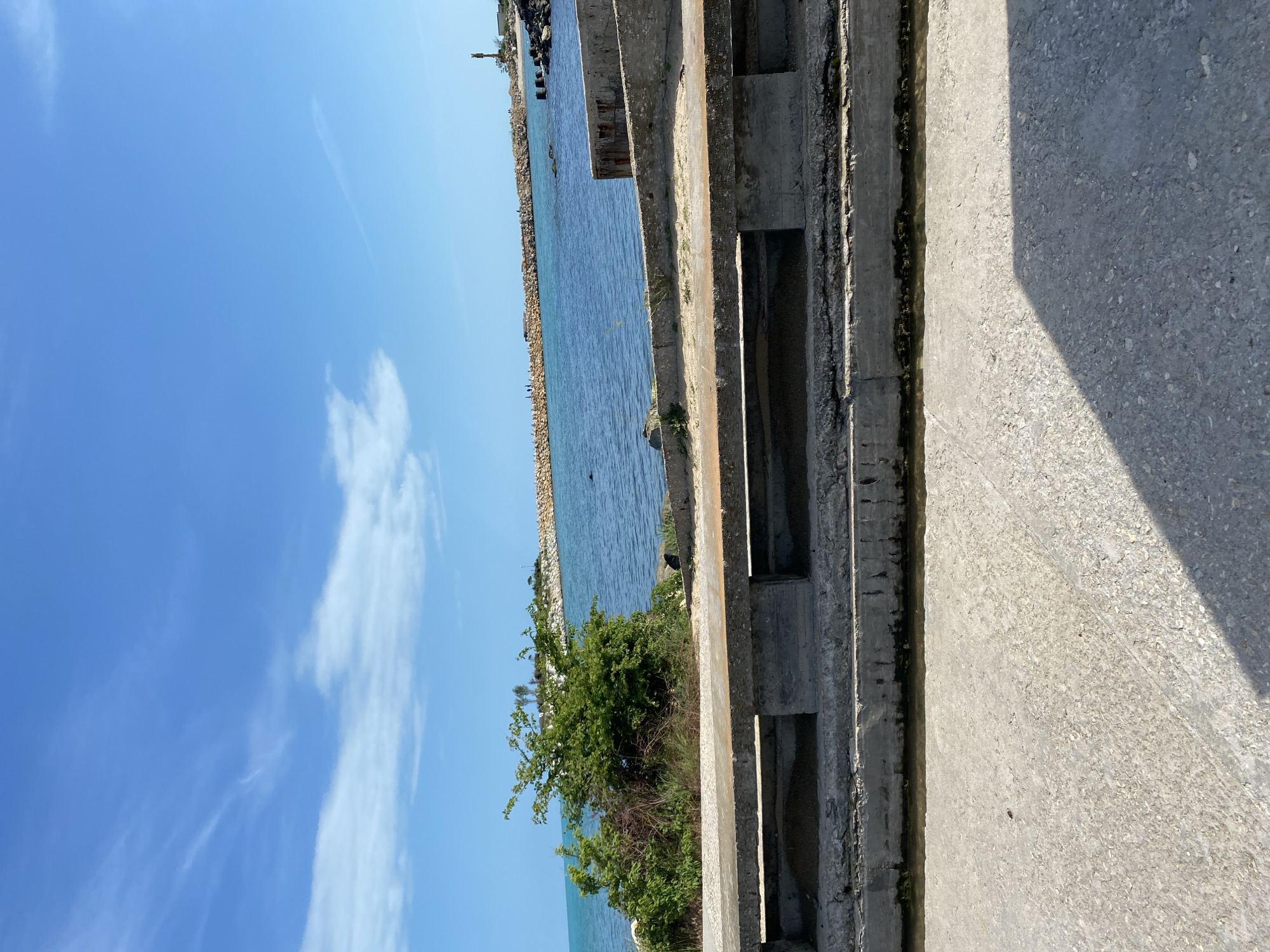 The image shows a coastal scene with a concrete structure in the foreground. The structure has rectangular openings and appears to be part of a pier or seawall. Beyond the structure, there is a body of water with a rocky shoreline. A small patch of vegetation grows near the structure. The sky is blue with scattered clouds. The location is Unknown, Unknown.