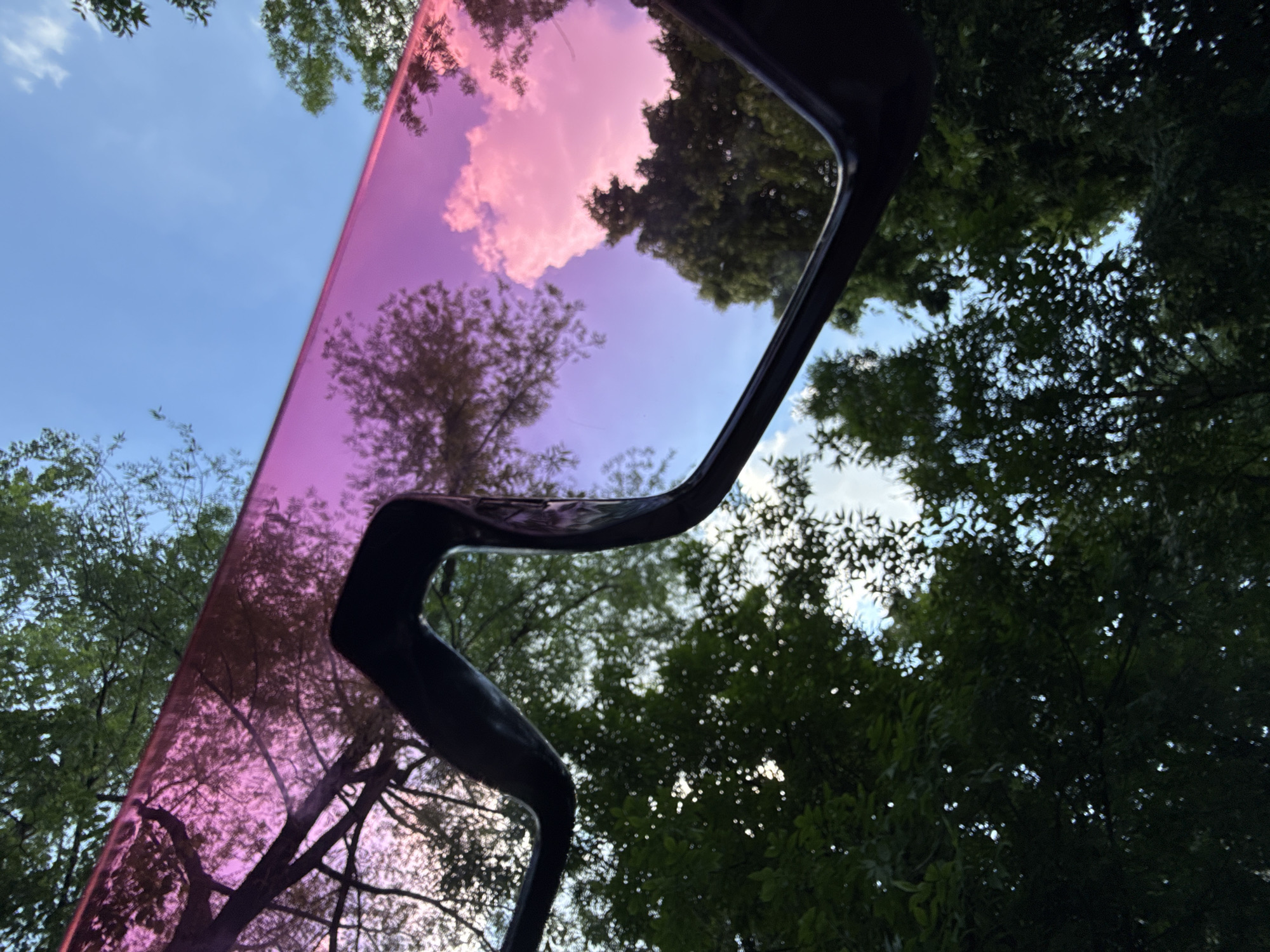 The image is a low-angle, close-up shot of a pair of pink-tinted sunglasses, seen from below. The sunglasses' frame is dark and wraps around the lens. The lenses reflect the sky, which is a bright blue with scattered white clouds. Pinkish clouds are also visible in the reflection, suggesting either a sunset or sunrise. The reflection also shows the silhouettes of tree branches and green foliage against the sky, indicating an outdoor, possibly wooded, environment. The lighting suggests it is daytime. No people, actions, or interactions are depicted. The image does not contain any information that would allow for its location to be identified as Moscow, Russia.