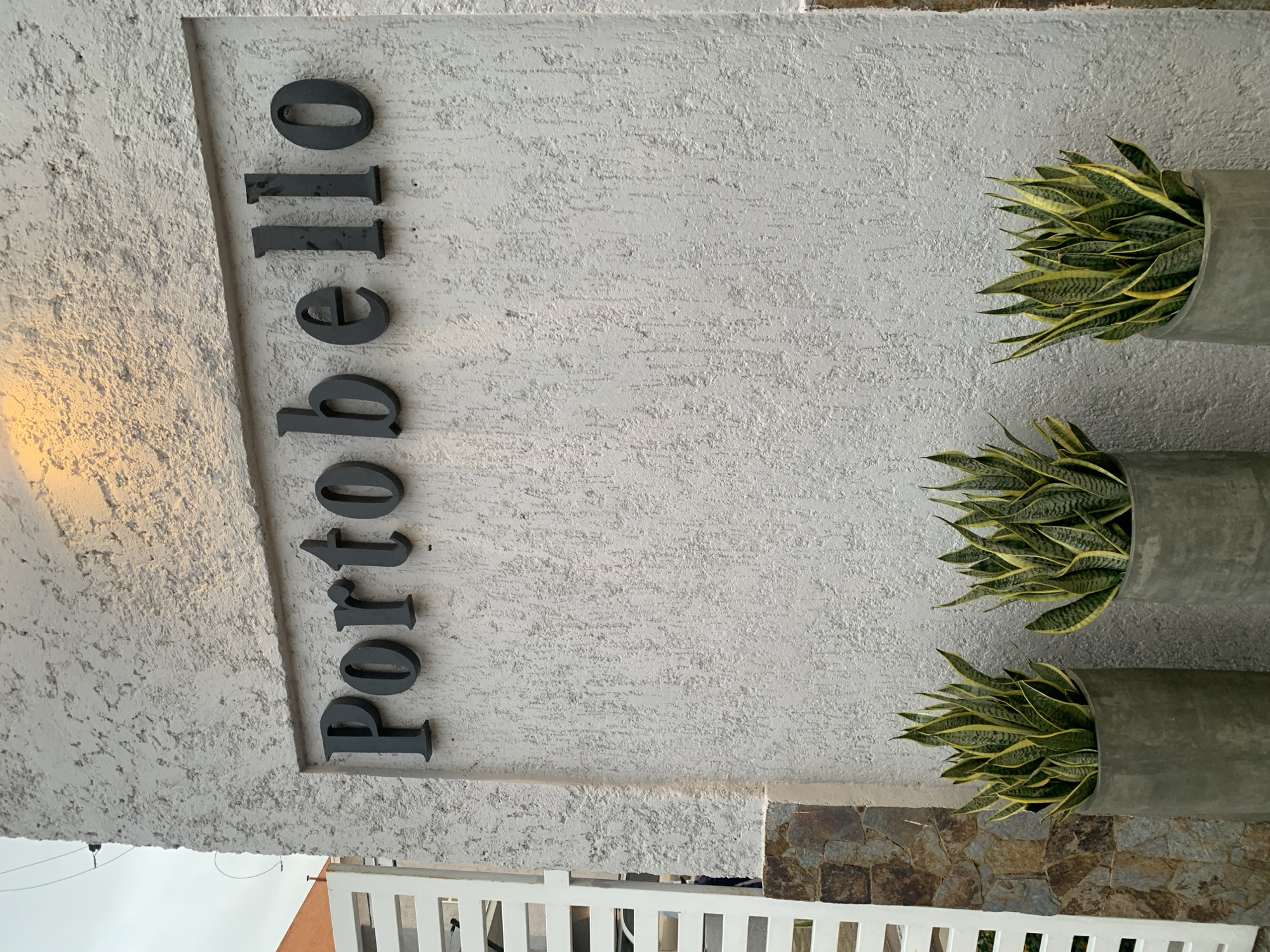 The image captures a detailed exterior view of a building facade, likely an entrance or a decorative wall section. The primary subject is a light grey, textured wall with the word "Portobello" spelled out in dark grey, raised, sans-serif letters, oriented vertically from top to bottom (or left to right if the image is viewed in a landscape orientation).
To the right of the text, three cylindrical concrete planters are mounted vertically along the wall. Each planter holds a dense cluster of variegated snake plants (Sansevieria trifasciata), characterized by their pointed, sword-like leaves with green centers and yellow margins. The plants appear healthy and well-maintained.
In the lower left portion of the image, a white, slatted fence is partially visible, hinting at a boundary or entrance. Below the main textured wall on the right, a section of a rustic stone wall or cladding is present, featuring irregularly shaped brown and grey natural stones. The sky, partially visible in the bottom left, appears overcast. A warm, bright light illuminates the upper left corner of the textured wall, possibly from a hidden light fixture or specific ambient light. The scene is static, with no people or activity depicted. The location context is Pampatar, Venezuela.