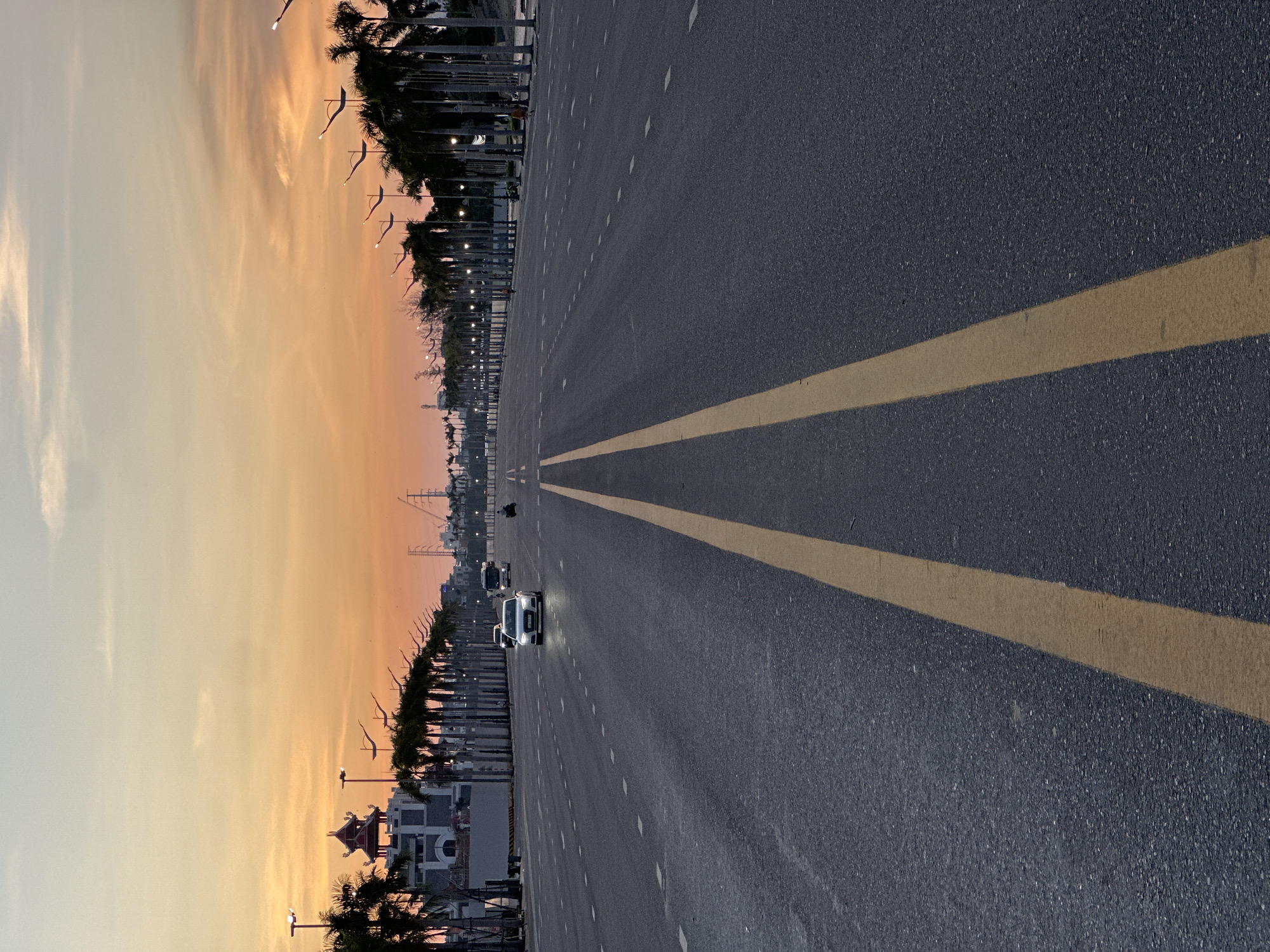 A wide, multi-lane road stretches into the distance under a colorful sunset sky in Da Nang, Vietnam. The road is paved with dark asphalt and marked with bright yellow and white lane dividers. On either side of the road are rows of tall palm trees, their fronds silhouetted against the orange and pink hues of the sky. To the left, a modern building with an Asian-inspired architectural element on its roof is visible, alongside streetlights and other buildings in the background. Several cars and a motorcycle are driving on the road. The overall impression is one of a peaceful evening scene on a well-maintained urban thoroughfare.