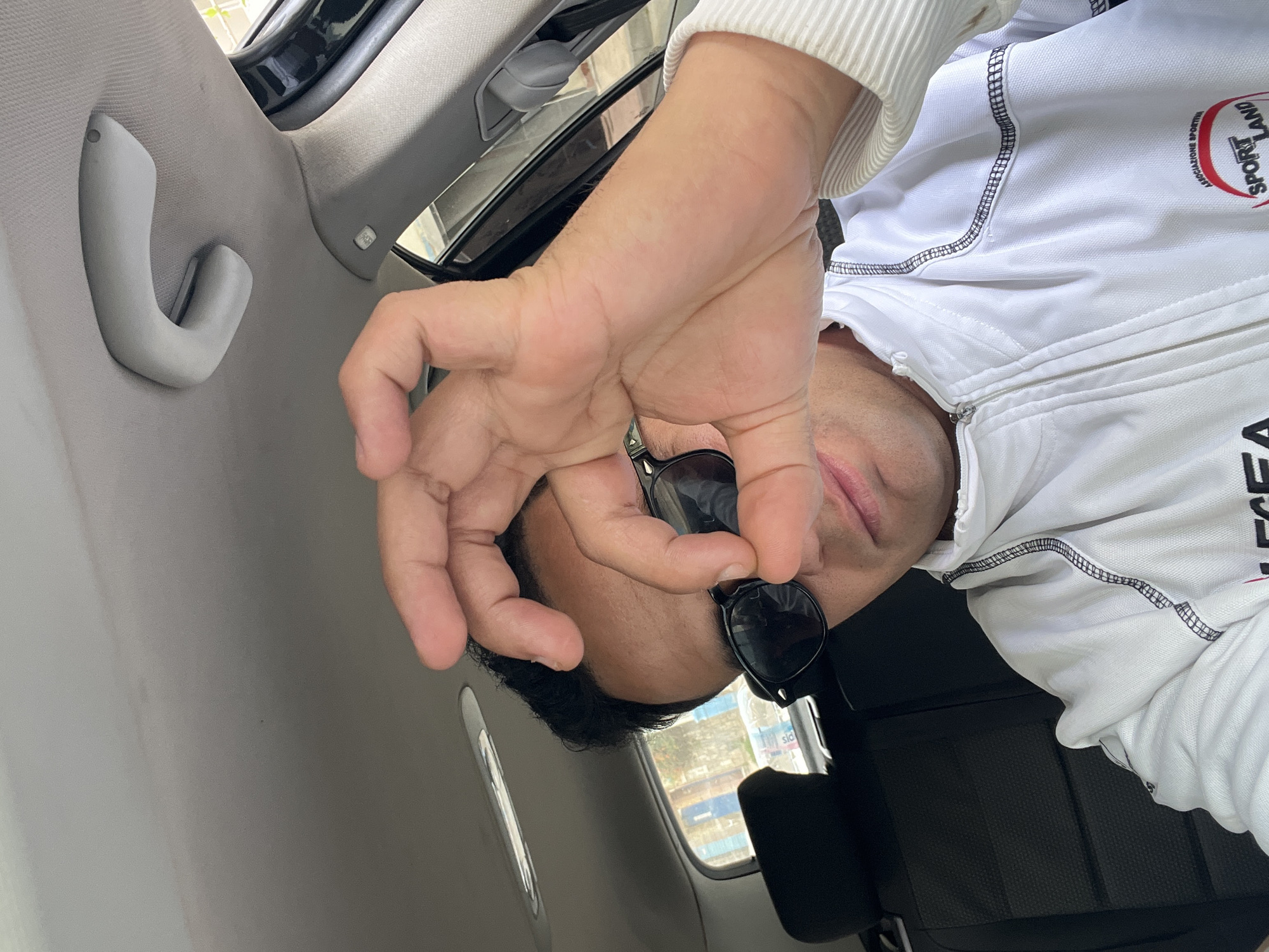 An upward-angled shot captures a person inside a vehicle, likely a car, in Martil, Morocco. The individual, appearing to be an adult male, is positioned on the right side of the frame with his head tilted. He is wearing sunglasses and a white zipped jacket adorned with black stitching. A red and black circular logo on the jacket's right chest features the text "SPORT LAND" and "ASSOCIATONE SPORTIVA" (Italian for Sports Association). The brand "LEGEA" is also partially visible on the jacket.
The person's right hand is prominently in the foreground, performing an "OK" gesture with the thumb and index finger forming a circle, while the other fingers are extended. His face is partially obscured by his hand and the sunglasses. The vehicle's interior features a gray fabric headliner and a curved gray grab handle. Through the window, daytime scenery is visible, including parts of buildings with blue accents and a water bottle with the letters "sic" visible on its label. A black seat is also discernible in the lower part of the frame. The overall impression is one of casualness.