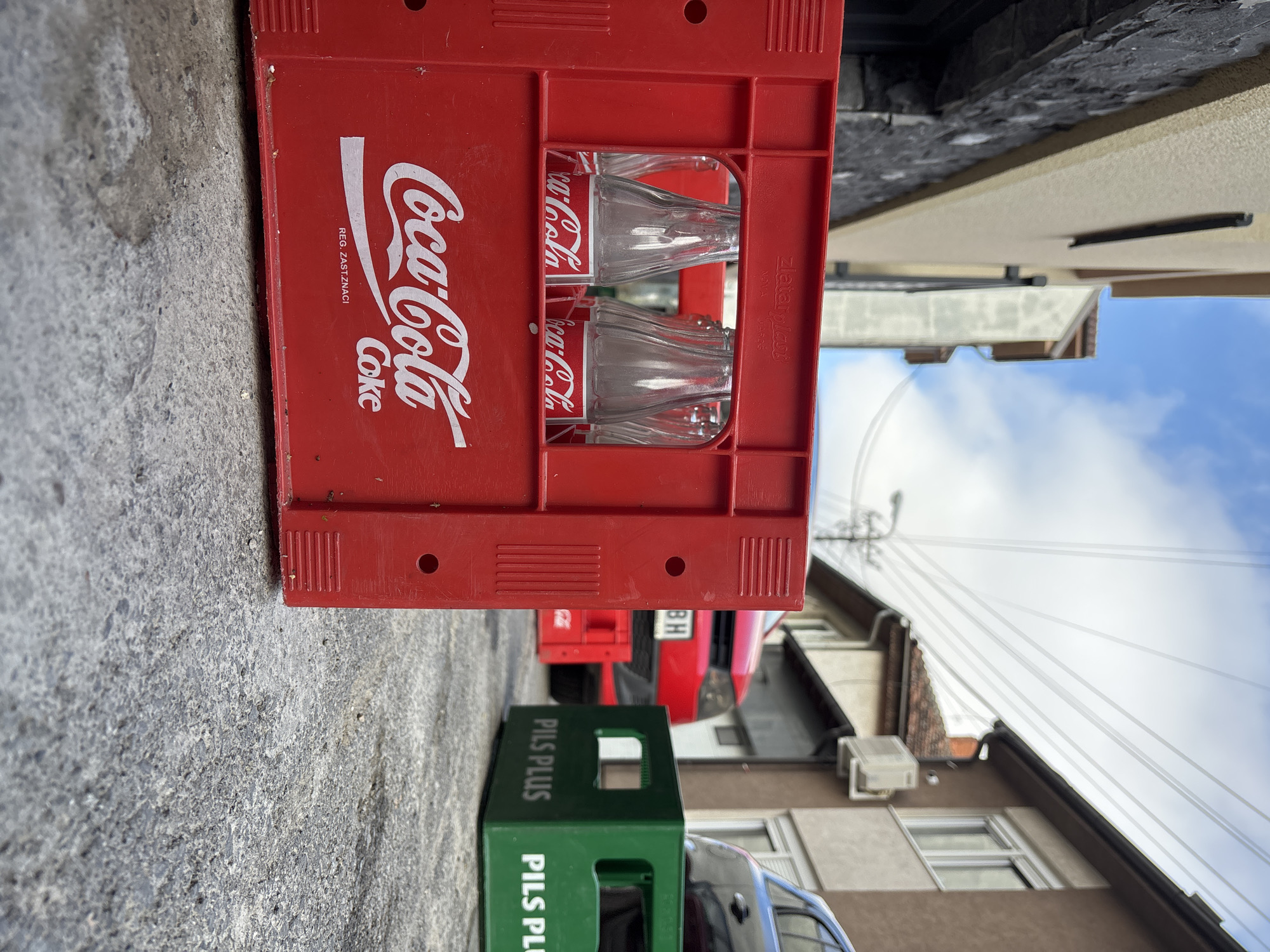 In an outdoor urban setting, a red Coca-Cola crate filled with empty glass bottles rests on a textured asphalt surface. The crate, featuring the iconic Coca-Cola logo in white, is positioned in the foreground and occupies the left half of the frame. Behind it, a green "PILS PLUS" crate is visible, along with a red vehicle and another dark-colored vehicle. The background shows buildings with windows and a cloudy sky with power lines overhead. The scene appears to be a street or alleyway, with no people or active interactions depicted.