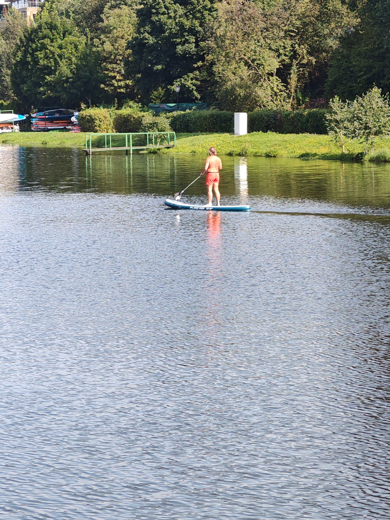 A lone individual is paddleboarding on a tranquil body of water. The person is wearing red shorts and is holding a paddle, actively engaged in the sport. The paddleboard is blue and white. The surrounding environment features lush green trees and vegetation along the shoreline, with some buildings visible in the background. A small green dock is also present near the shore. The water exhibits gentle ripples, reflecting the surrounding greenery and the sky. The image appears to be taken during daylight hours, suggesting a pleasant outdoor setting. This scene could plausibly be located in the city of Rosinka, Russia, given the natural elements and potential for recreational water activities.