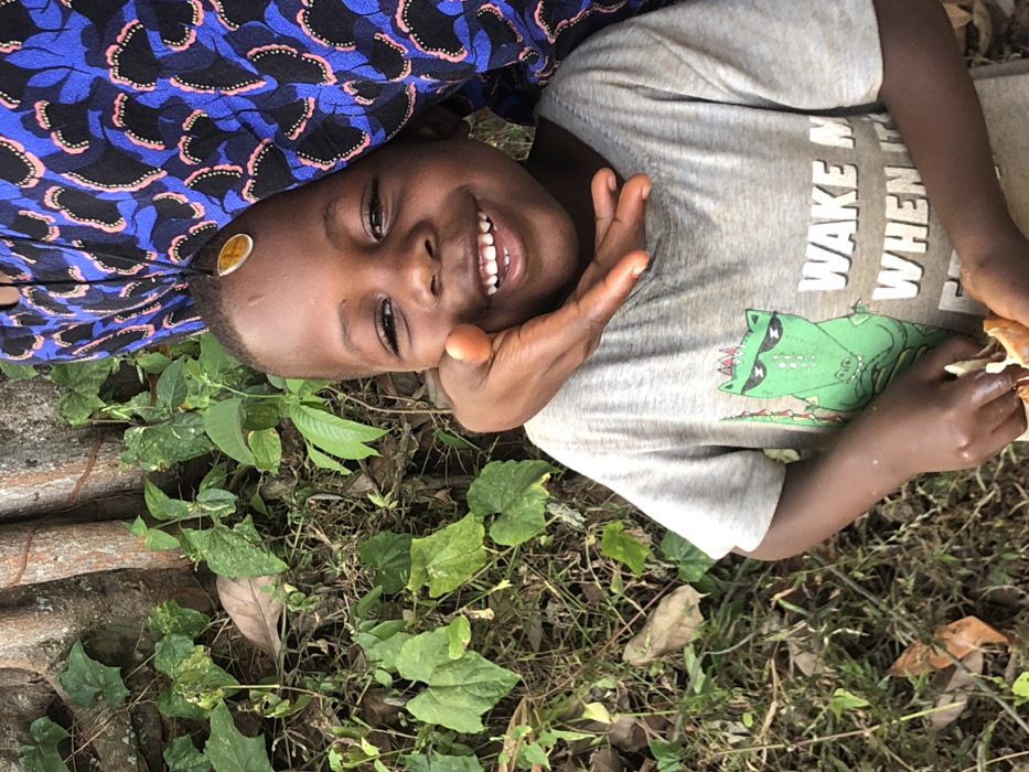 A smiling child lies on the ground amid greenery. The child wears a gray t-shirt with a green alligator graphic and the words "WAKE ME UP WHEN IT'S." A hand rests near the child's face. A blue and black patterned fabric is visible near the child's head. A yellow object is stuck on the child's forehead. The background includes grass, leaves, and tree roots. The image is taken at an angle, rotated slightly.