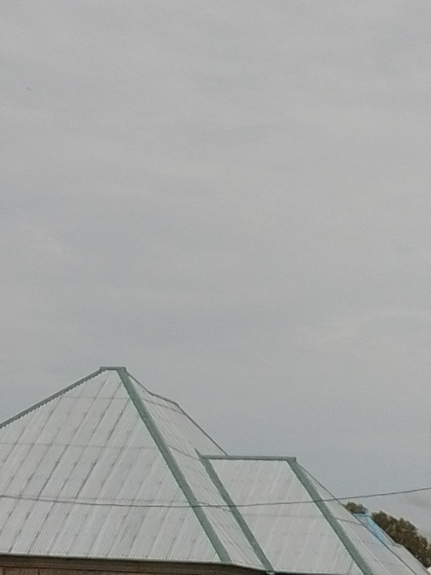 The image shows a portion of two corrugated metal roofs, one larger than the other and in the foreground, meeting at a ridge. The roofing material is light gray and appears somewhat weathered, with visible horizontal corrugations. Green trim lines the edges of the larger roof. The sky above is overcast and a pale gray, with no discernable features other than a faint dark speck, possibly a distant bird or debris. The lower portion of a wooden structure is visible beneath the larger roof. In the background, on the right side of the image, the tops of green trees are visible, suggesting an outdoor setting. There are no people or specific actions depicted. The scene appears to be a quiet, overcast day in a location that could be within the city of Howayi, Nigeria, characterized by the typical architecture of such regions.