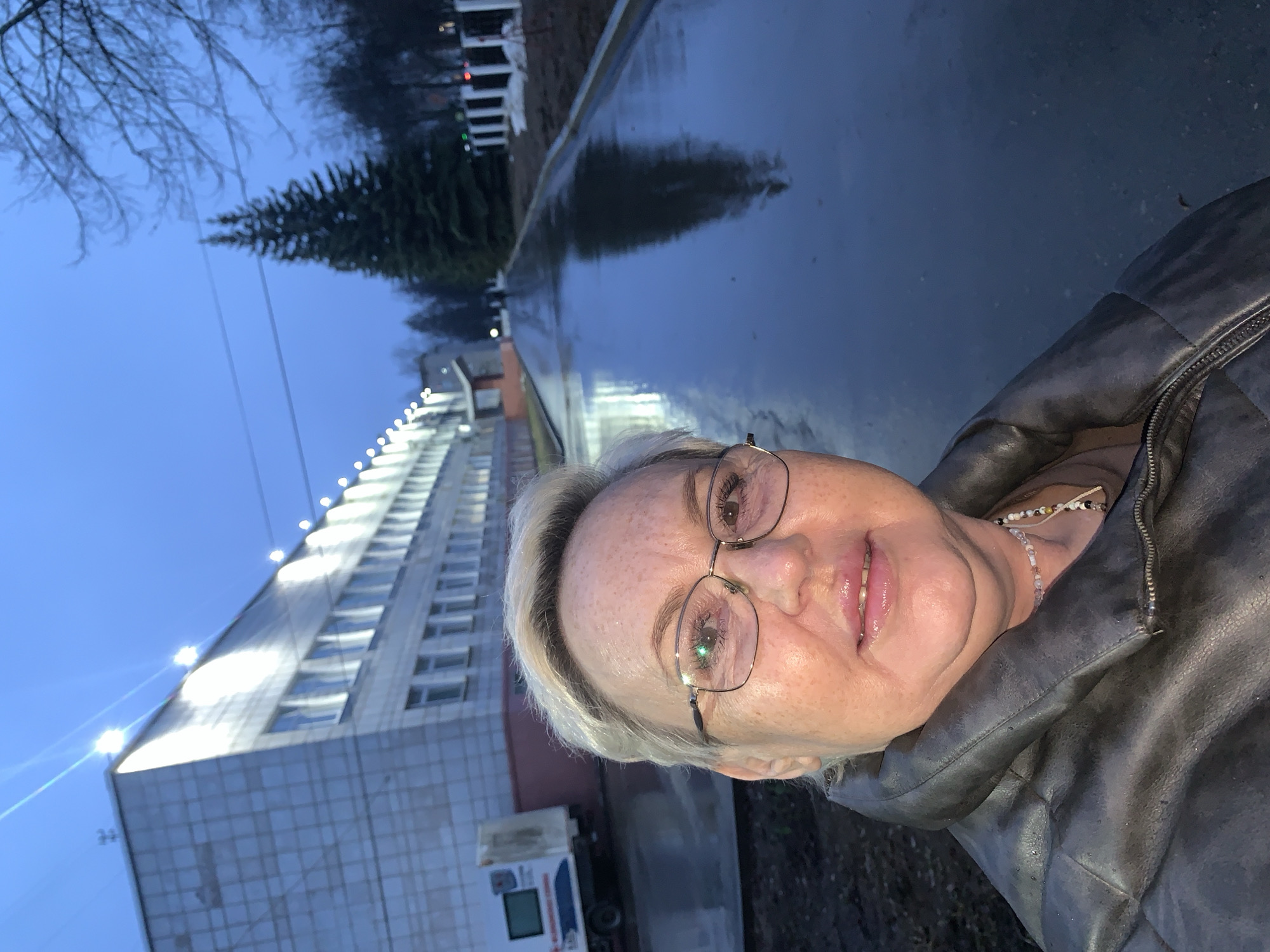 A woman with short blonde hair and glasses is taking a selfie. She is wearing a dark leather jacket and a beaded necklace is visible. The background is a wet, paved area, suggesting it has recently rained. A multi-story building with rows of windows is visible on the left, illuminated by a series of lights along its upper edge. In the distance, a pine tree and other buildings are present. The scene appears to be in an urban setting at dusk or night. Based on the location metadata, the city is Chaikovsky, Russia.