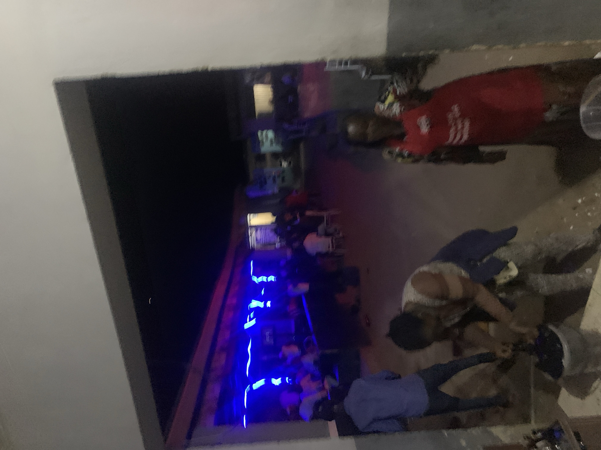 The image, captured at night in New Stadium, Nigeria, displays an outdoor or semi-outdoor social venue. In the foreground, a male figure in a blue long-sleeved shirt and dark trousers stands with his back to the camera on the left. Centrally, a female individual, wearing a light-colored patterned top and dark lower garments, is bent over a dark bucket, appearing to mix its contents with an implement. Another dark bucket sits beside it. To the right, another female individual stands, generally facing the camera, wearing a red apron with white, partially illegible text over a multi-colored patterned shirt. The ground in the foreground is a dark, uneven surface, likely compacted earth or concrete, with sparse light-colored debris.
The midground features an illuminated section of the venue, distinguished by horizontal blue and vertical purple neon strip lighting affixed to an overhead structure. Numerous blurred individuals are visible within this area, some seated at tables and others standing, suggesting a social gathering. The background consists of dark, indistinct building structures under the night sky.