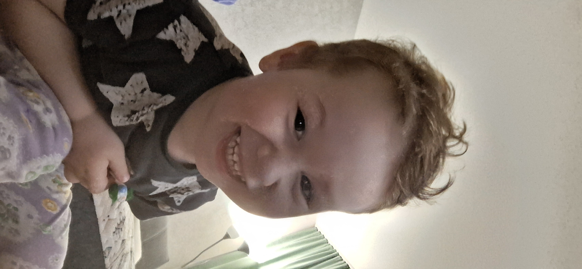 A young child with light brown curly hair is smiling and looking at the camera. The child is wearing a dark t-shirt with white star patterns and is holding a small object in their left hand. The background appears to be a bedroom setting with bed linens visible on the left and a light-colored wall on the right. The location is unknown, but given the request, it is assumed to be the city of Chaikovsky, Russia.