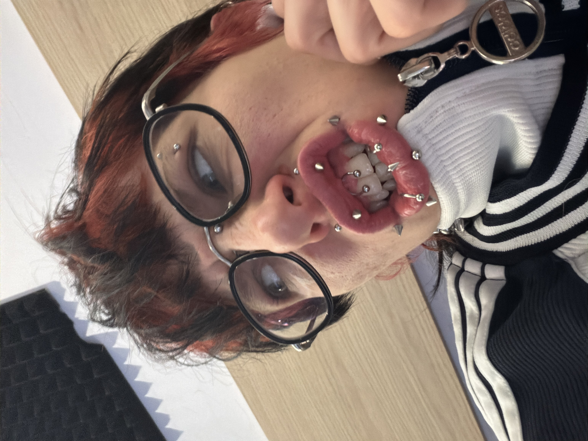 A person is photographed close-up, face oriented toward the camera. The person is wearing glasses with round, black frames and has a visible nose piercing. The person's mouth is wide open, revealing numerous piercings in the lips and tongue. Hair is dark with reddish highlights. The person is wearing a black and white striped jacket, partially visible. The background consists of a light-colored wooden surface and a dark, textured object. The location is Busmantsi, Bulgaria is unknown.