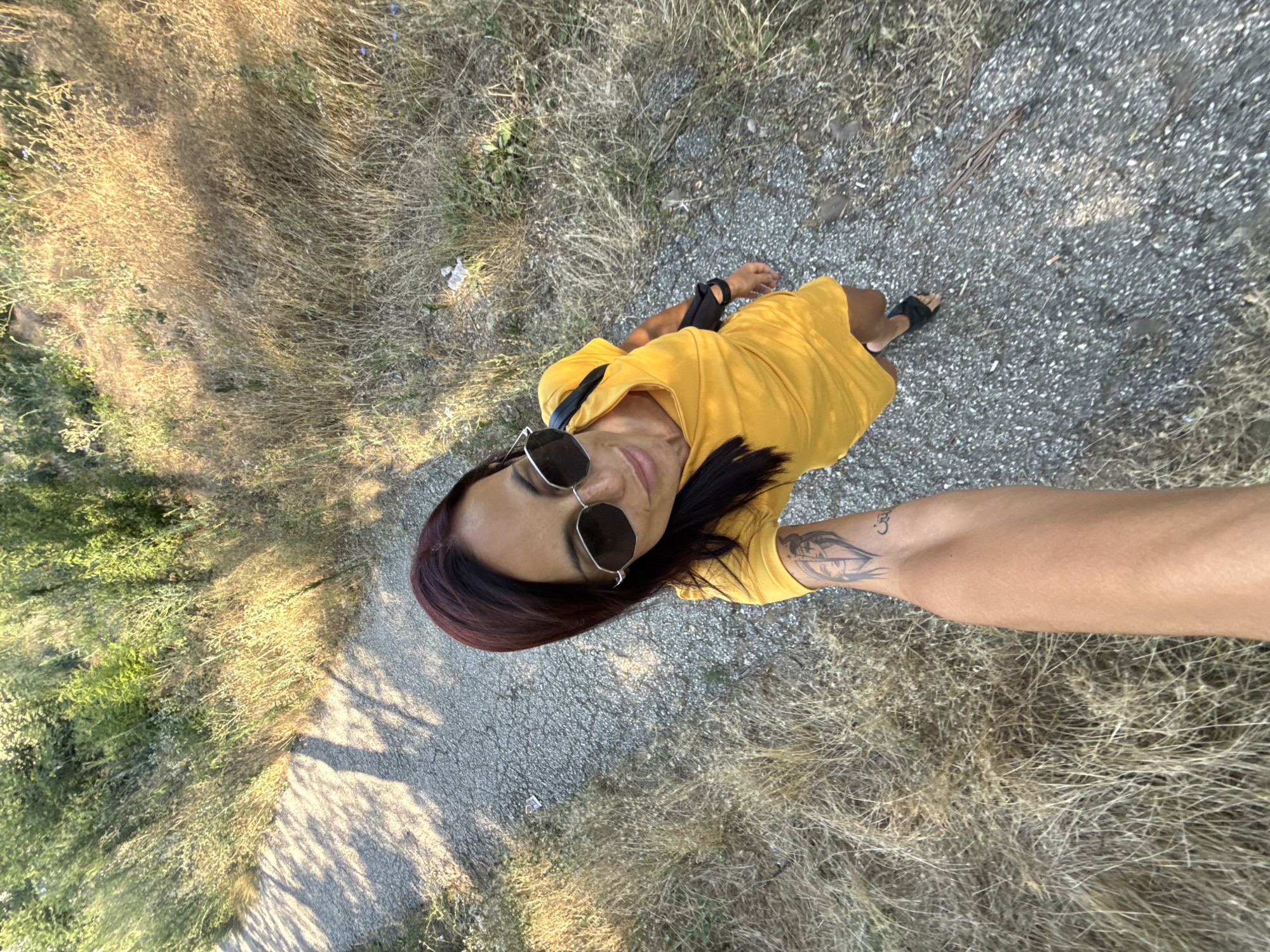 In Pleven, Bulgaria, a woman with dark hair and sunglasses is captured in a high-angle selfie. She wears a bright yellow dress and black sandals. Her left arm, visible in the foreground, displays a tattoo. The scene unfolds on a gravel path bordered by dry, golden grasses and green foliage, under natural daylight casting shadows.