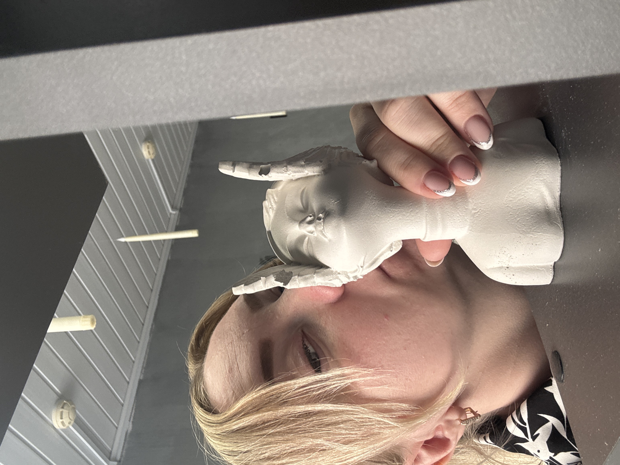 A woman with blond hair is holding a white, plaster-like object shaped like an elephant's head up to her face. Her left hand is grasping the elephant's head. She is wearing a black and white patterned top, and a gold earring is visible. The background appears to be an indoor space with gray, striped paneling and visible lighting fixtures. A dark gray surface is present in the foreground. The image does not have any geographic coordinates; the location in the city of Chaikovsky, Russia, is not supported.