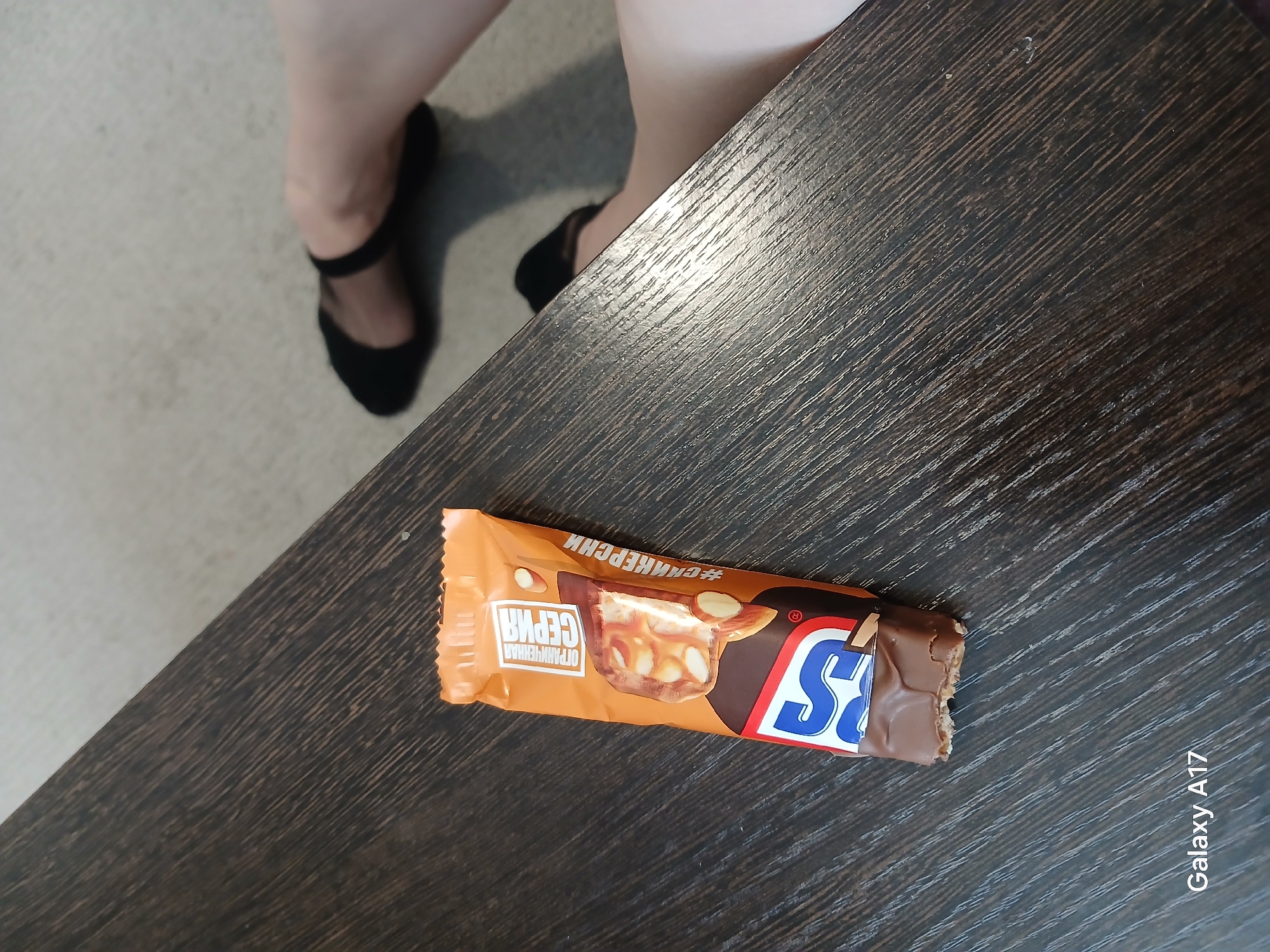 The image depicts a partially unwrapped Snickers bar on a dark brown, textured surface resembling a wooden tabletop. The Snickers wrapper is orange with Russian text. A pair of legs wearing black sheer tights and black shoes are visible in the background, to the left of the table. The image includes the watermark "Galaxy A17". Based on the Russian text on the Snickers wrapper, this image was likely taken in Russia. Assuming the phone's location settings were on when the photo was taken, it is possible it was taken in Yekaterinburg. However, this cannot be confirmed from the image itself.