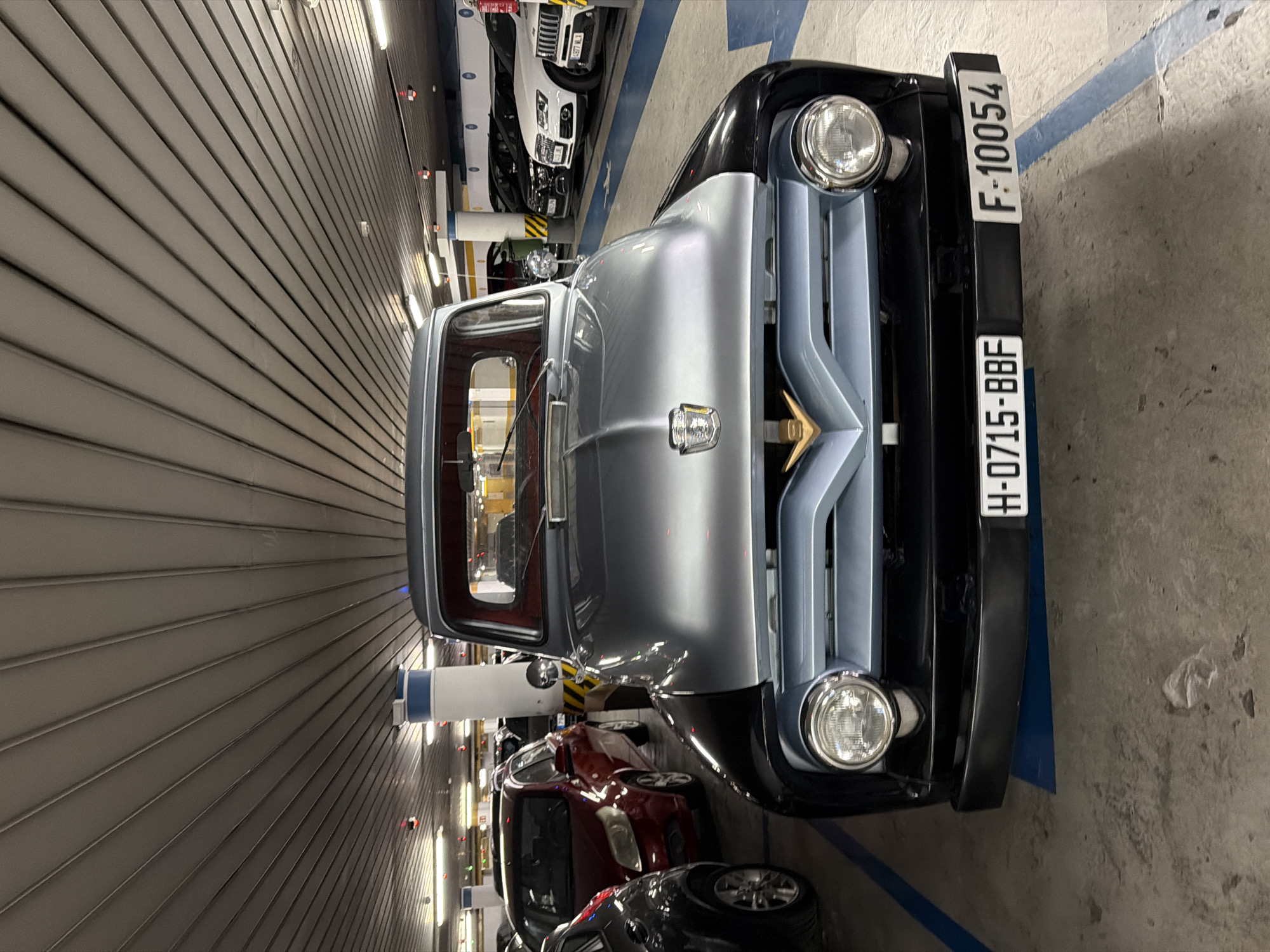 The image shows a vintage Ford pickup truck parked in a multi-story parking garage. The truck is silver and black with a light blue grill. It has round headlights and a gold emblem on the hood. The license plate reads "H-0715-BBF F-10054". Other vehicles, including a red hatchback and several white SUVs, are visible in the background. The parking garage has a sloped ceiling with fluorescent lighting and blue painted lines on the floor marking parking spaces. There is a white pillar with blue and yellow stripes to the right of the truck. The scene appears to be static, with no people or actions depicted. The location is identified as Benidorm, Spain, though this cannot be determined from the visual evidence alone.