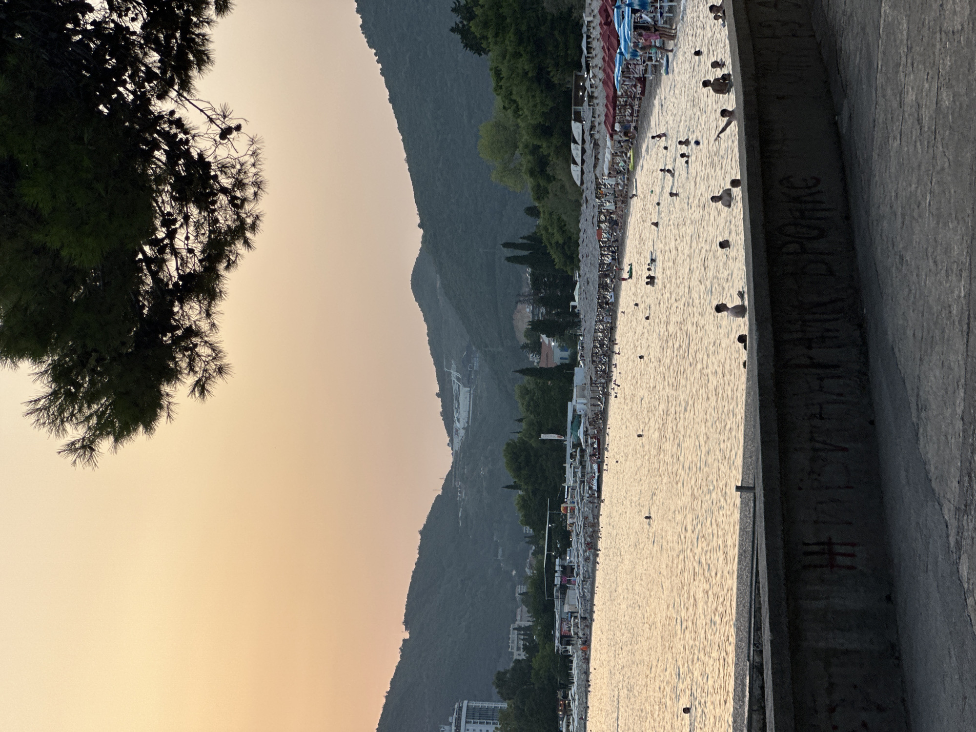 A wide shot captures a beach scene in Budva, Montenegro, during the golden hour. The sky is a gradient of soft orange and pale yellow. In the foreground, a concrete barrier is visible with graffiti scrawled across it in red and white. Beyond the barrier, a calm, sun-drenched sea reflects the warm light, with numerous people scattered throughout the water, some swimming and others wading. The beach is lined with rows of straw umbrellas and lounge chairs, occupied by many sunbathers. Behind the beach, lush green hills rise up, dotted with trees and buildings, including a tall white hotel. The overall impression is one of a serene and lively evening at the coast.