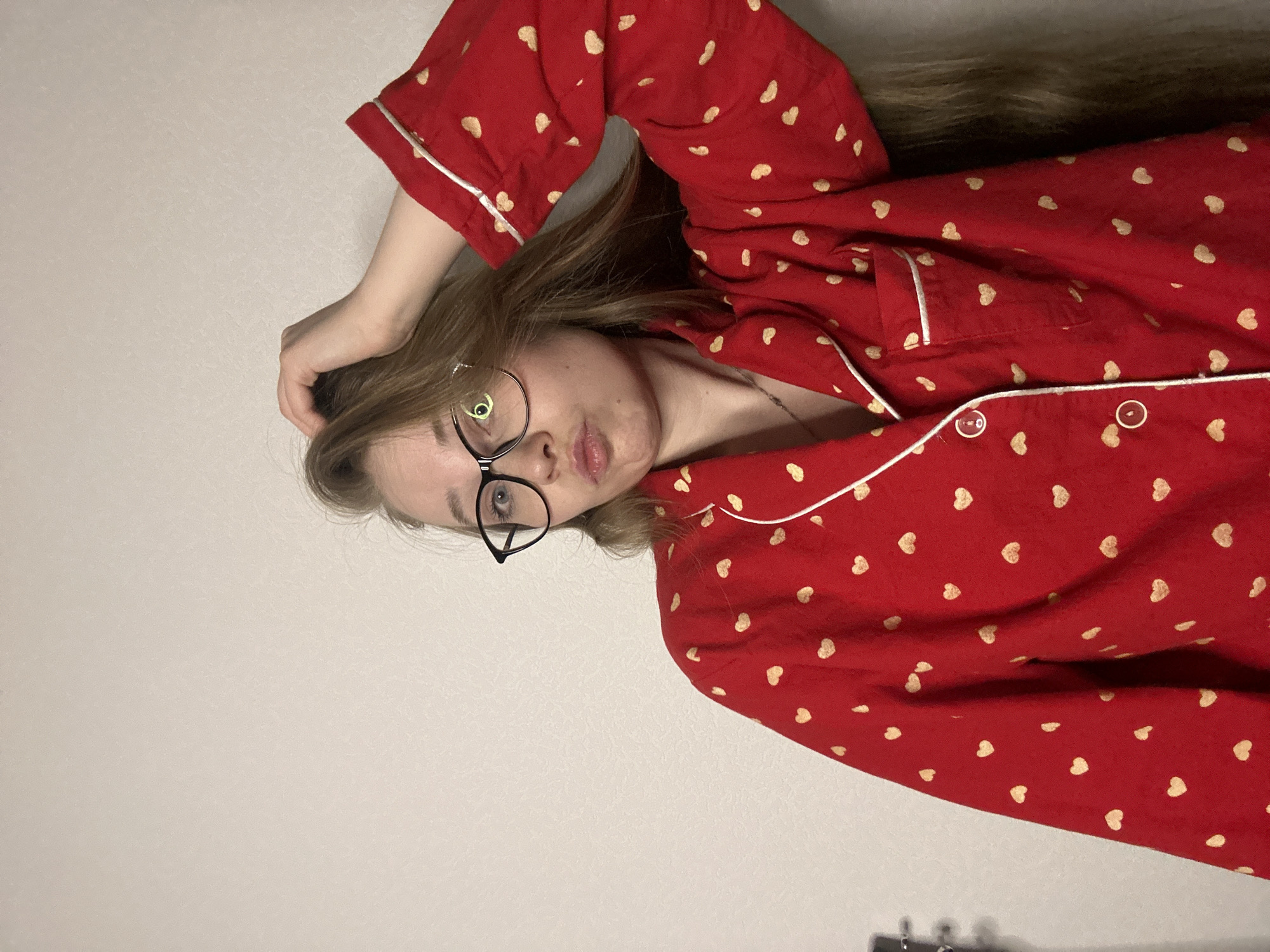 A young woman with light-colored hair and black-framed glasses is depicted lying down, possibly on a bed or sofa. She is wearing a red pajama top adorned with numerous small, golden heart patterns and white piping along the edges and pockets. Two white buttons are visible on the front of her top. Her left arm is bent with her hand resting near her head, and her hair is spread out behind her. She has a slight pout on her lips and is looking directly at the camera. A thin necklace with a small pendant is visible around her neck. The background is a plain, light-colored wall, possibly off-white or light gray. The image is oriented sideways, with the woman's head to the left and her body extending to the right.