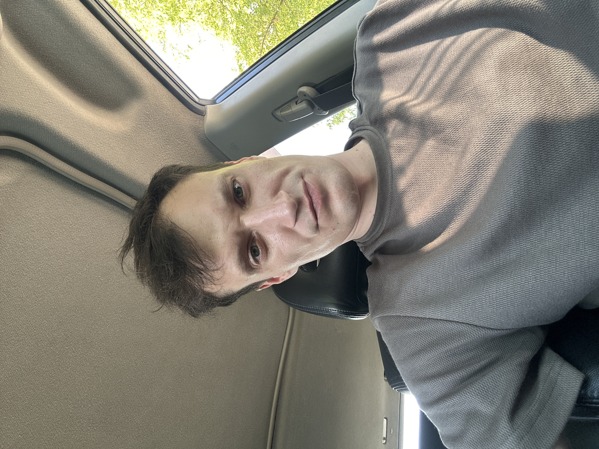 This is a selfie taken inside a car. The person in the image is a white male with short, dark brown hair and fair skin. He is wearing a grey t-shirt. He is looking directly at the camera with a neutral expression. The car's interior is visible, including the headrest of the seat and the car's ceiling. Sunlight is streaming in from the right side of the car, casting shadows on the person's face and t-shirt. Outside the car window, green trees are visible, suggesting an outdoor location in a temperate climate, possibly during daytime. The image does not provide specific details about the city of Makiv, Ukraine, or its surroundings.