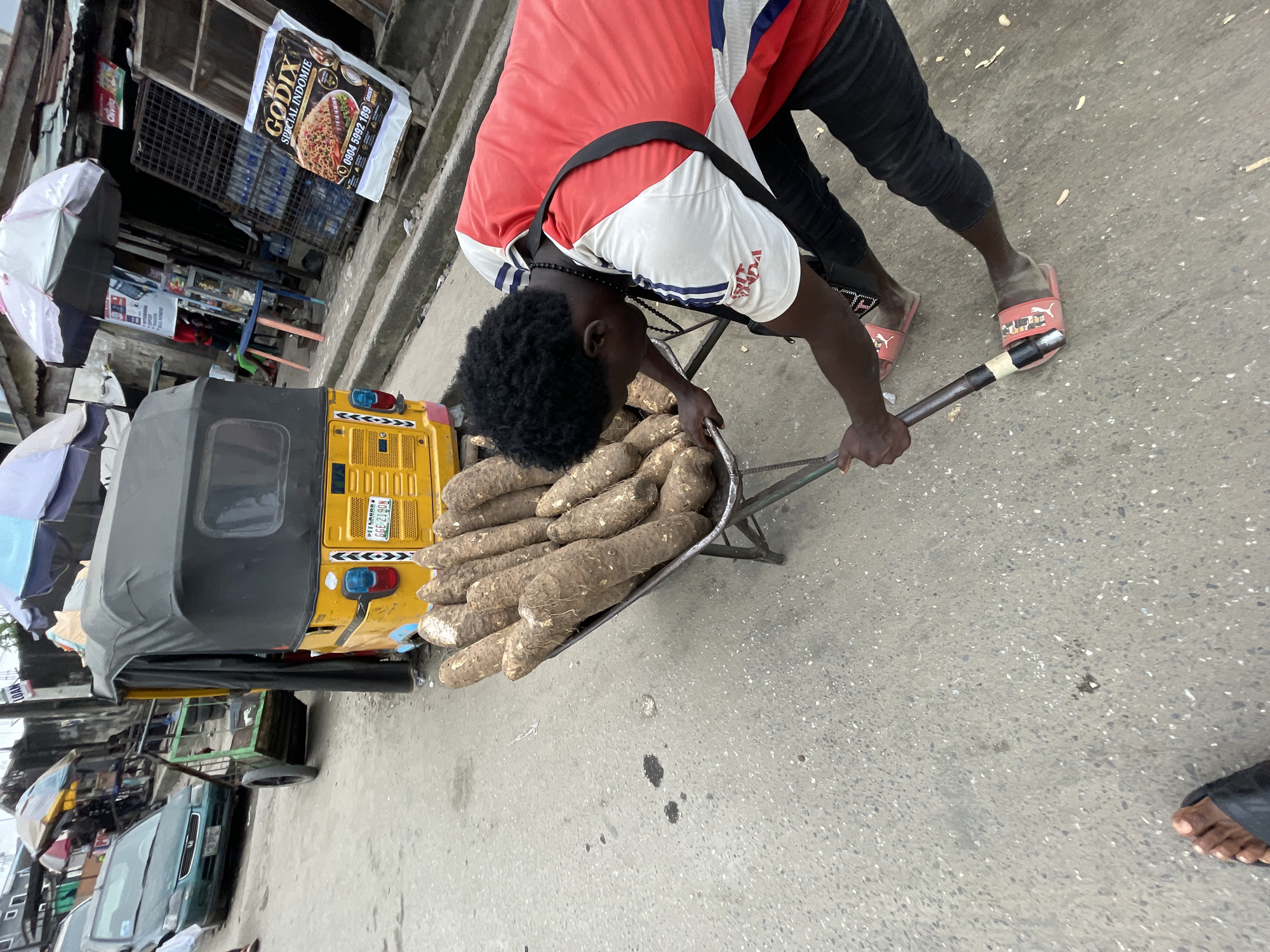 In Ado, Nigeria, on a paved street, a person is pushing a wheelbarrow loaded with large yams. The wheelbarrow's handles are wrapped in black tape. The person, wearing a red and white t-shirt and black pants, is bent over, pushing the wheelbarrow with their right hand. They are wearing pink slides with a white logo. Behind the wheelbarrow, a yellow auto-rickshaw with a black canopy is parked. The street is lined with stalls and shops, some with awnings and umbrellas. A green car is visible in the background to the left, and a rack of bottled water is stacked in front of a shop to the right. A flyer advertising "GODIX SPECIAL INDOMIE" is prominently displayed on the right side of the image. The environment suggests a busy market or commercial area.