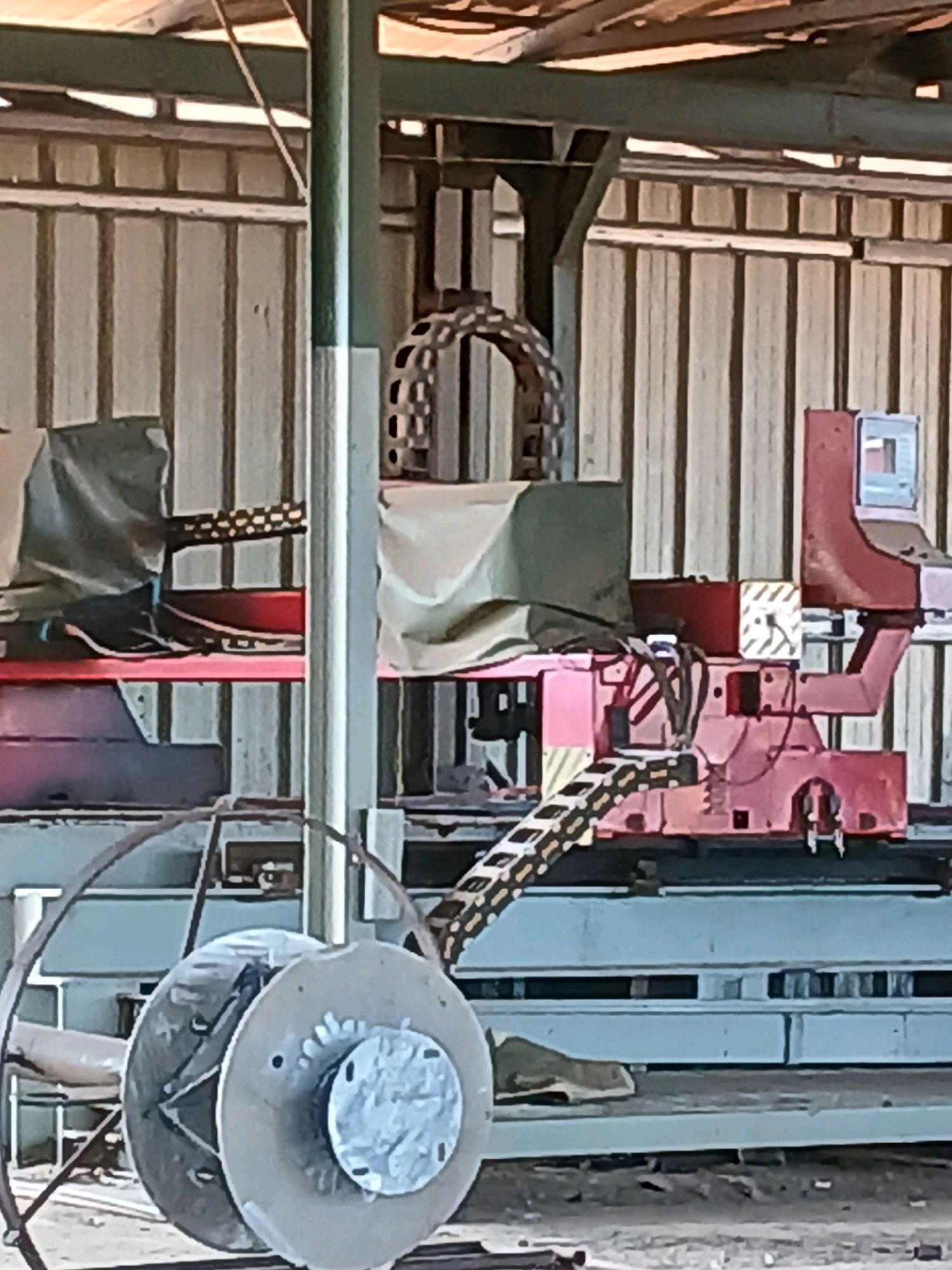 The image depicts an industrial setting, possibly a workshop or a factory, located in Pangkalpinang, Indonesia. The scene is characterized by heavy machinery and industrial equipment. In the foreground, a large, circular object, resembling a spool or reel, is prominently visible, mounted on a frame. Behind it, a complex piece of machinery with red and pink components is in operation. This machinery features a conveyor belt system with patterned links, suggesting it is involved in material handling or processing. A monitor or screen is attached to one part of the machinery, indicating a control or monitoring station. Various metallic structures and supports are visible throughout the scene, contributing to the industrial aesthetic. The background consists of corrugated metal walls and a partially visible roof structure, further emphasizing the industrial environment. The overall impression is one of an active industrial workspace in Pangkalpinang.