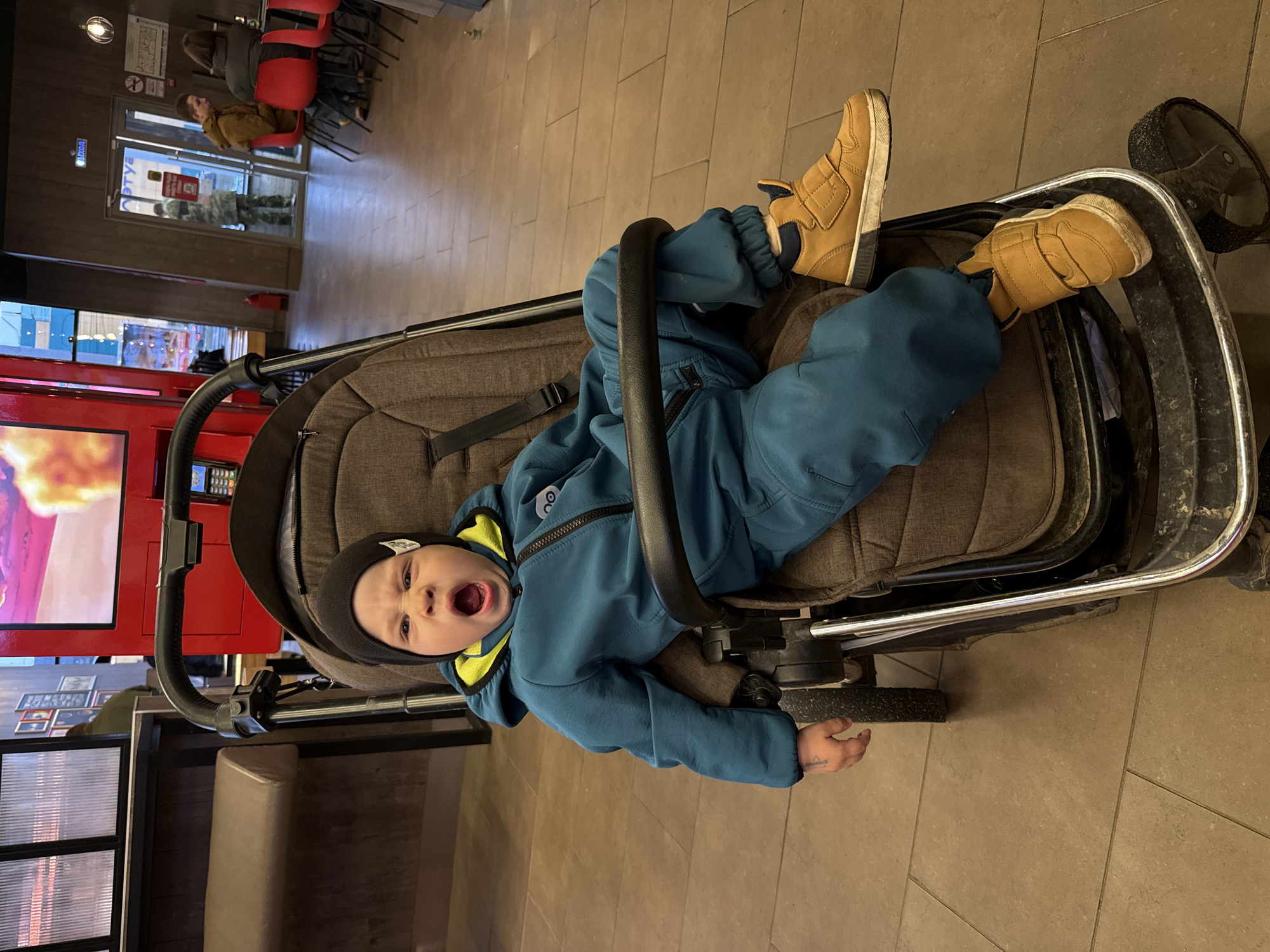 A young child wearing a blue snowsuit and brown shoes sits in a stroller in what appears to be an indoor dining area. The child is wearing a dark beanie and has their mouth open, possibly yawning or shouting. The stroller is positioned on a light brown tiled floor. In the background, there are red chairs and a glimpse of an exterior with people walking by. A red vending machine-like structure is also visible in the background. The location is identified as Rusanovo, Russia.