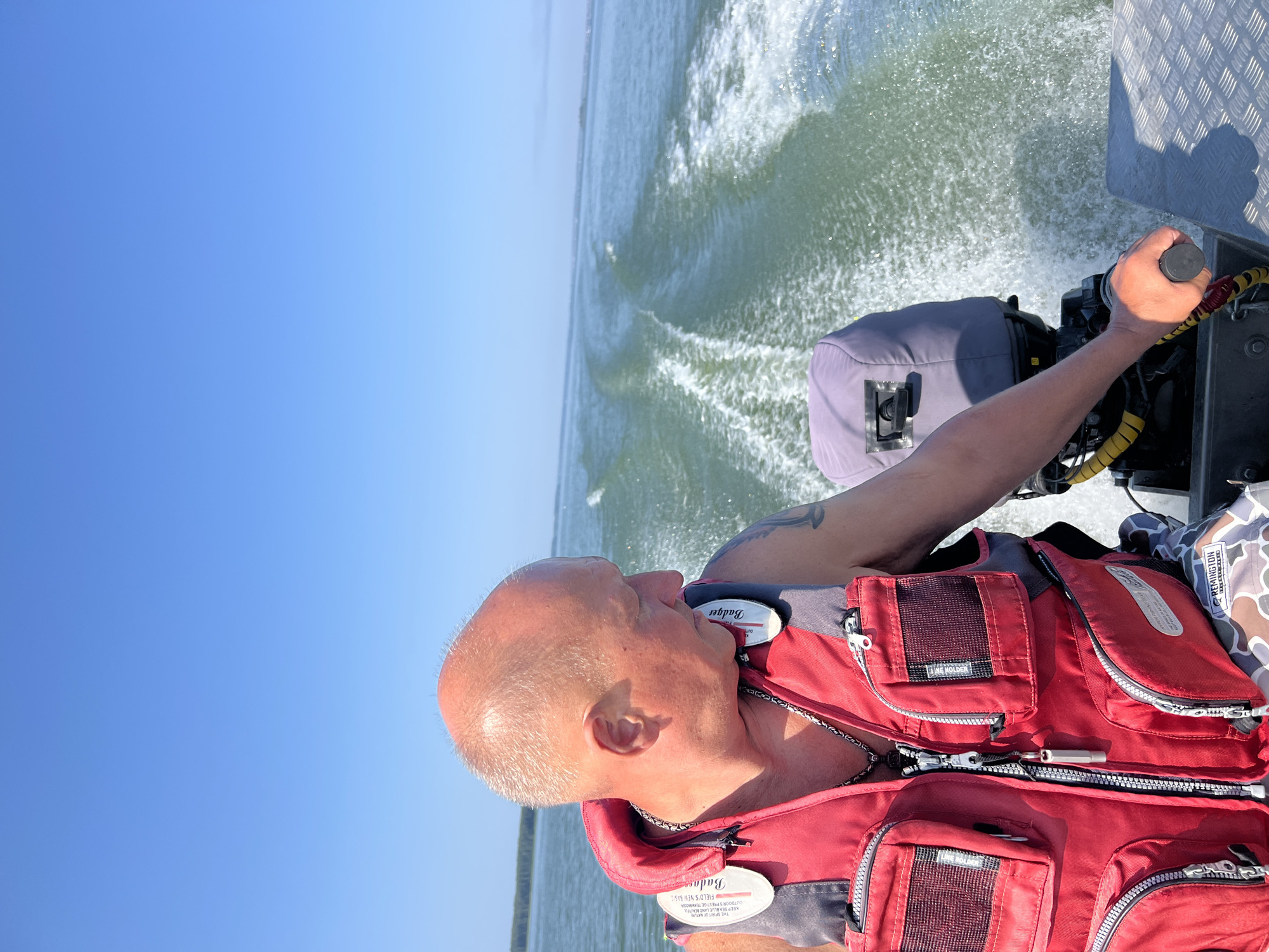 A man is operating a motorboat on a clear day on the sea. The man is wearing a red life jacket and a silver chain necklace. The boat is moving at a good speed, creating a wake in the water. The water is a light green color, and the sky is a clear blue. There is a line of trees in the distance on the horizon. The scene appears to be taking place in Beregovoe, Russia, as indicated by the text on the life jacket.