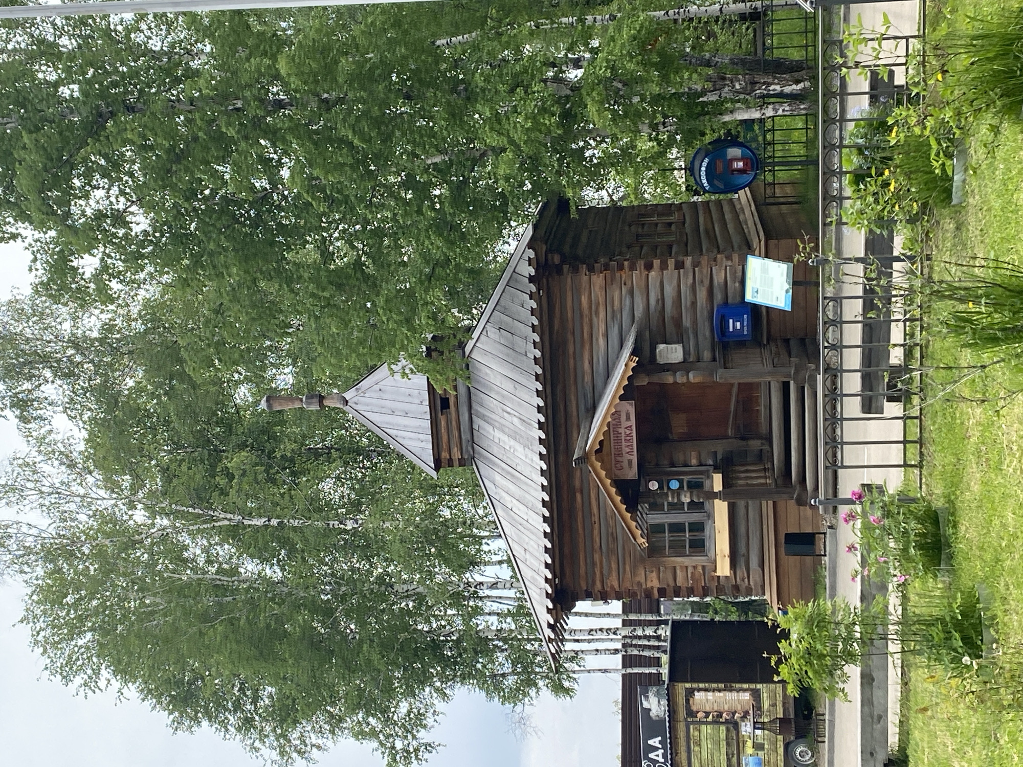 The image shows a traditional Russian wooden building with a tiered roof and a small porch. The building is constructed from logs and features small windows. Above the entrance, a sign with Cyrillic text, possibly indicating a shop or service, is visible. To the left of the wooden building, there is a food truck or trailer with green and yellow branding. In the foreground, a black metal fence encloses a grassy area with some flowering plants. Behind the fence, a blue public telephone booth with Cyrillic text is mounted on a pole, and a blue mailbox is attached to the wooden building. The scene is set against a backdrop of tall birch trees with green foliage, suggesting a natural or park-like environment. The sky is overcast. The location is identified as Тальцы (иркутский), Russia.