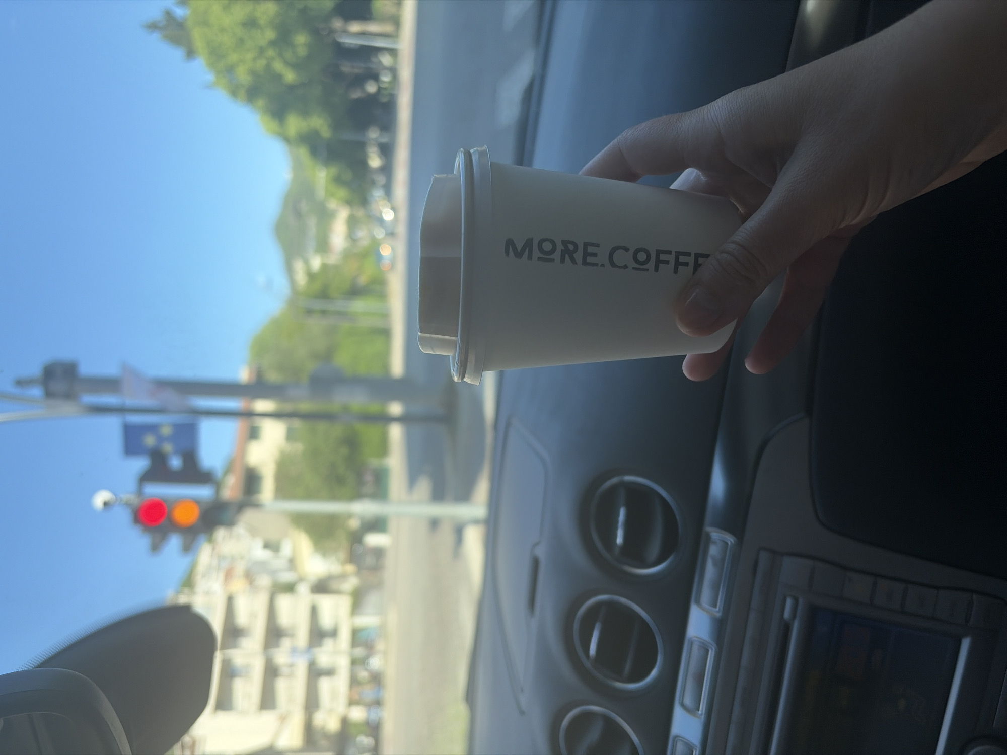 The image shows a person sitting in a car, holding a cup of coffee with the words "MORE.COFFEE" on it. The scene outside the car window is blurred, but appears to be a street in a city. A traffic light is visible, showing red and yellow lights. The city is likely Budva, Montenegro, given the presence of the traffic light and a hint of the urban landscape.