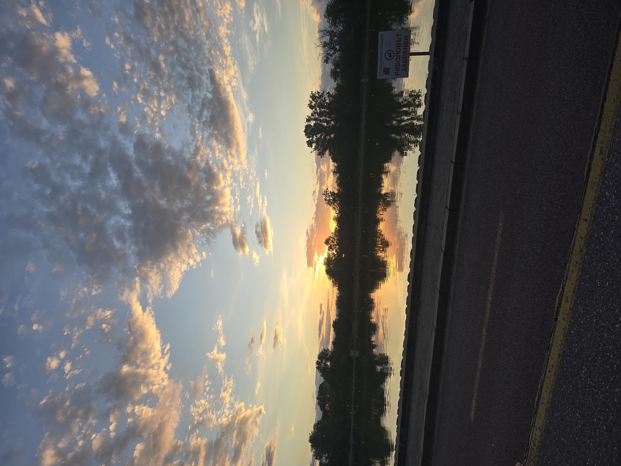 The image depicts a sunset reflected in water, likely a canal or river, in the city of Plovdiv, Bulgaria, as suggested by the sign that says "ГРЕБЕН КАНАЛ ПЛОВДИВ". The clouds in the sky are dappled with shades of white, gray, and gold from the setting sun. Trees line the edge of the water, their silhouettes and reflections creating a symmetrical composition. A paved area with a yellow line marking is visible along the water's edge. The sky is a mix of blue and orange, transitioning from light to dark.