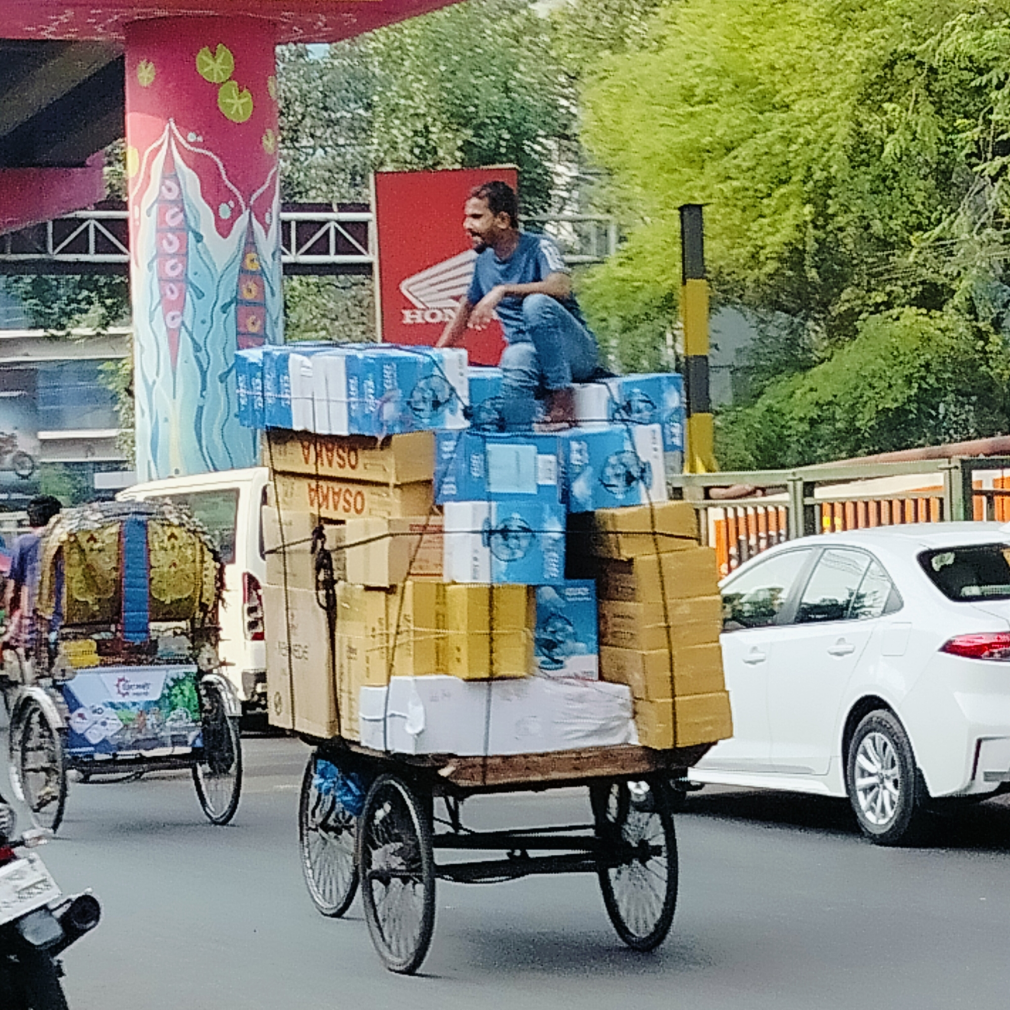 This daytime image captures a vibrant street scene in Dhaka, Bangladesh, centered around a man transporting goods. A man with a beard, wearing a blue t-shirt and jeans, sits on top of a dangerously high stack of packages loaded onto a three-wheeled cargo bicycle. The cargo consists of numerous brown cardboard boxes and blue-and-white bundles, some labeled "OSAKA" (visible upside down). The man appears relaxed, showing a slight smile.
To the left, a traditional pedal-powered rickshaw, adorned with colorful decorations, moves along the street. A white modern sedan is partially visible on the right side of the frame. In the background, lush green trees and bushes indicate a leafy urban environment. Architectural features include a large, red pillar decorated with distinct patterns resembling lotus leaves or water lilies, suggesting artistic civic infrastructure. A red billboard with the "HONDA" logo is also visible behind the man, alongside a black and yellow striped pole. The scene portrays the bustling daily life and methods of goods transportation in a South Asian city.