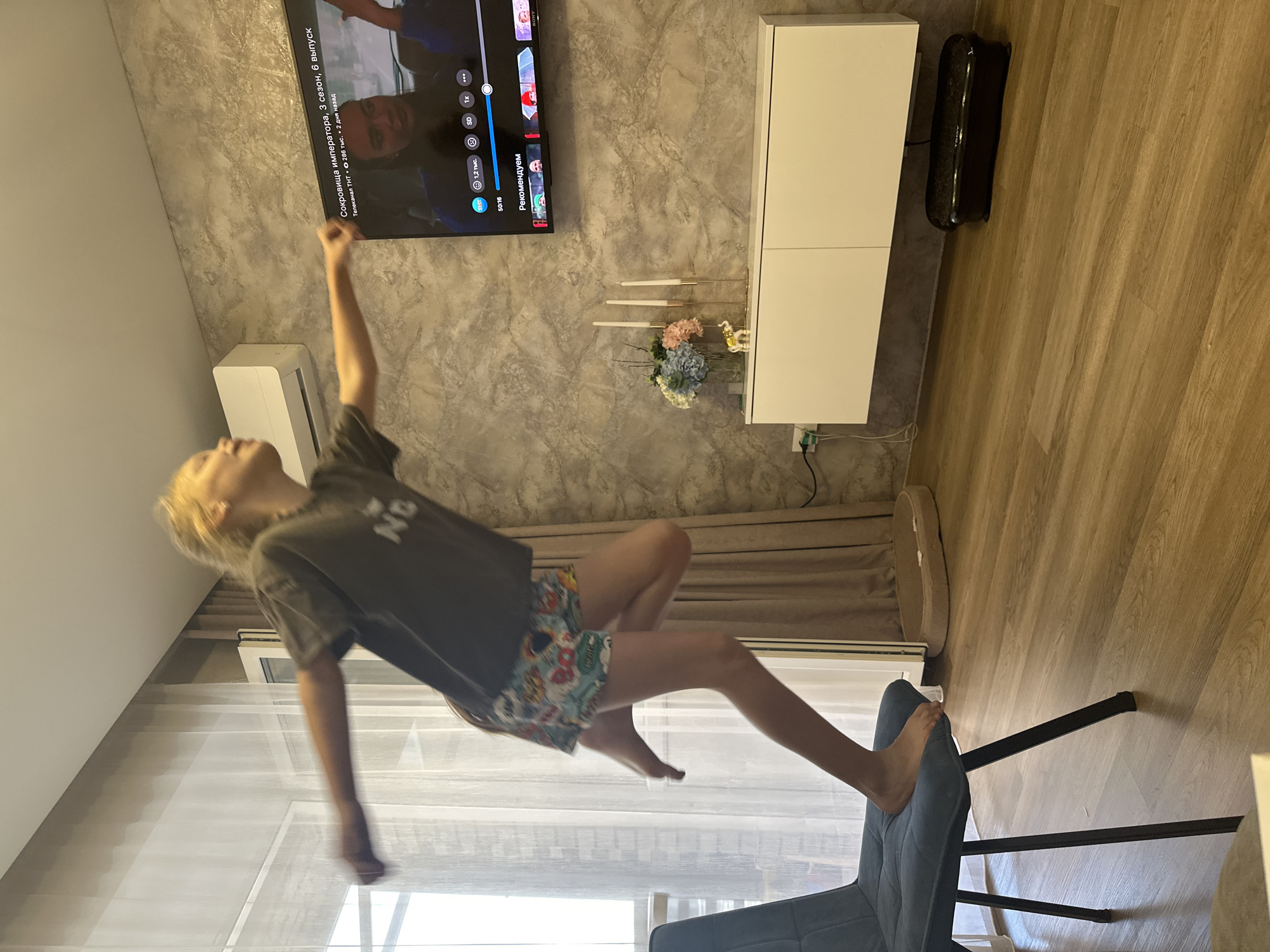 A young person with blonde hair, wearing a dark t-shirt and patterned shorts, is captured mid-air, seemingly jumping or falling. Their body is angled downwards, with legs bent and arms outstretched. The person is in focus, appearing somewhat blurred due to motion.
The scene takes place indoors, likely a living room. A large television screen hangs on a textured, gray wall, displaying a show with Russian text, including "Сокровища императора" (Treasures of the Emperor), "3 сезон, 6 выпуск" (Season 3, Episode 6), and "Телеканал ТНТ" (TNT Channel). A white air conditioning unit is mounted below the TV.
To the right of the TV is a white cabinet with two doors. On top of the cabinet are three tall, thin candles in holders, and a small bouquet of artificial flowers in blue, pink, and white. A small golden figurine of a dog is also on the cabinet. A black object, possibly a soundbar or a sub-woofer, is placed on the wooden floor next to the cabinet.
The floor is made of light brown wooden planks. A gray upholstered chair is partially visible in the foreground, with the person's foot resting on its back. A white sheer curtain is visible behind the person, suggesting a window or patio door. The lighting appears to be artificial, casting a warm glow on the wooden floor. The overall impression is a candid shot of a playful moment at home.