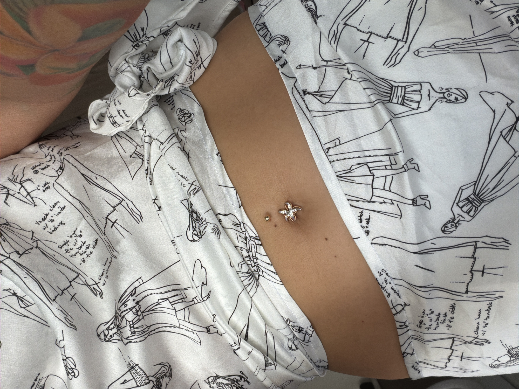The image shows a close-up of a person's midriff with a belly button piercing. The person is wearing a white garment with black line drawings of fashion sketches and technical sewing notes. The piercing is a rose gold color with a star-shaped charm adorned with clear stones on the bottom, and a small gold ball on the top. The background is out of focus. There is no indication of location in Nha Trang Ward, Vietnam.