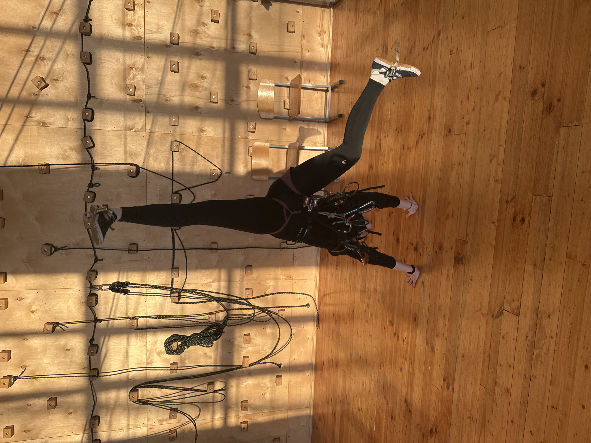 A woman in black athletic leggings, a dark top, and athletic shoes is suspended horizontally between a plywood climbing wall on the left and a wooden plank floor on the right. She is secured by a climbing harness and numerous ropes attached to wooden climbing holds on the wall. Her legs are split, one extending towards the climbing wall and the other towards the wooden floor. Her arms are outstretched, one hand near the climbing wall and the other near the floor. In the background, two wooden school chairs are partially visible. The lighting is uneven, casting shadows across the climbing wall and the floor. The location is Chaikovsky, Russia.