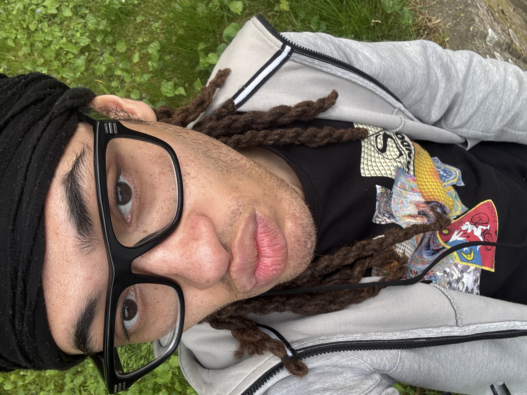 The image shows a person lying on grass. The person has brown dreadlocks and is wearing a black head wrap. They have black-framed glasses on their face. The person is wearing a black t-shirt with a colorful graphic print and a gray zip-up hoodie. The background consists of green grass, suggesting an outdoor setting. There is no other information available to identify the location as Les Ulis, France.