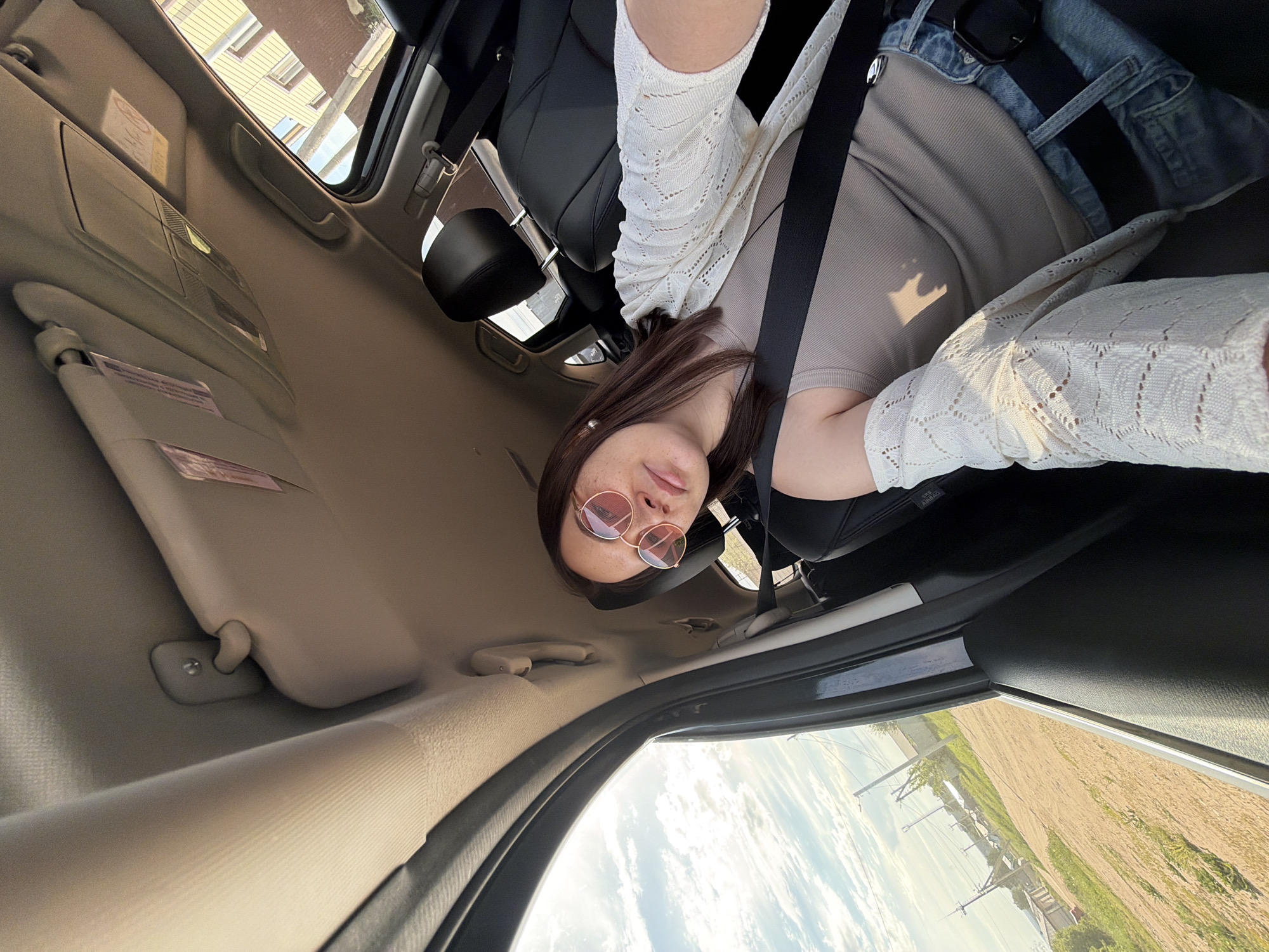 A person is taking a selfie inside a car, wearing round rose-tinted glasses, a beige top, a white crocheted cardigan, and a denim jacket. They are wearing a seatbelt. The interior of the car is visible, including the sun visor and sunroof. Outside the window, the landscape suggests a rural or semi-rural area with fields, power lines, and buildings in the distance. While the location is stated as Chaikovsky, Russia, the image itself does not provide explicit markers or signage to confirm the precise location.