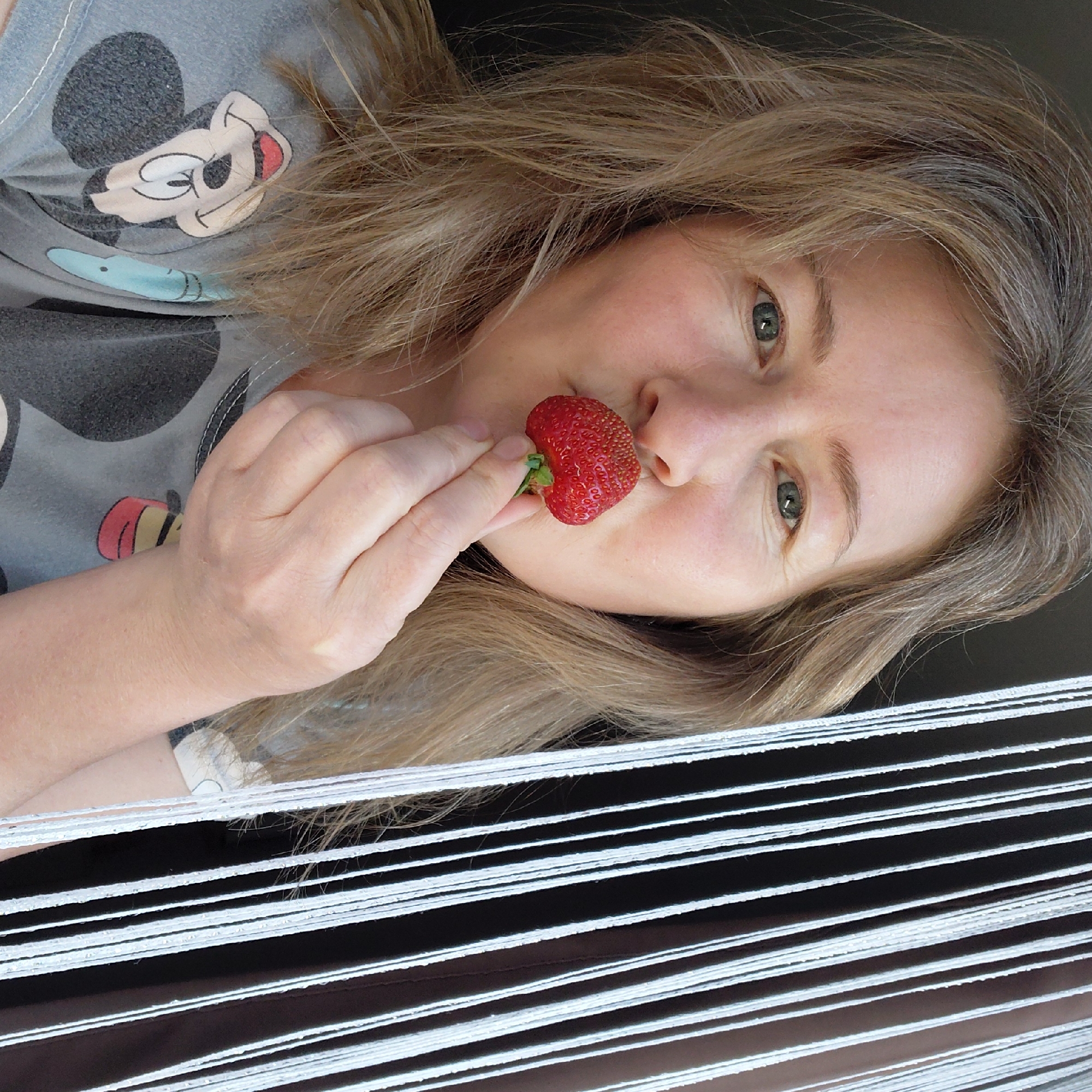 A medium shot captures a fair-skinned Caucasian woman in her late 30s or early 40s holding a ripe strawberry to her lips. She has shoulder-length blonde hair with streaks of grey, and her eyes are a hazel green. She is wearing a grey t-shirt with a cartoon print of Mickey Mouse. The background is out of focus, featuring vertical white strings on the right side and a dark area on the left. The overall lighting suggests an indoor setting with natural light filtering from the side. The image does not contain any identifiable landmarks or features that would indicate its location in Novosibirsk, Russia.