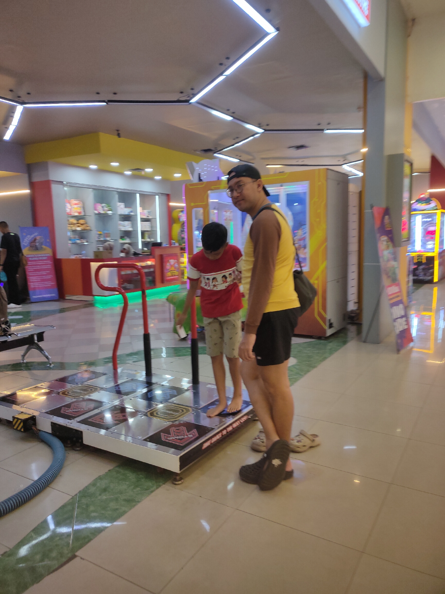 The image depicts an indoor arcade or entertainment center, likely located in Kediri City, Indonesia, as suggested by the contextual information. The scene is illuminated by fluorescent lighting and decorative strip lights on the ceiling. In the foreground, a dance game machine with a grid of illuminated pads is visible. A young boy wearing a red t-shirt with a graphic print and patterned shorts is standing on the dance pads, barefoot. To his right, an adult male wearing a yellow tank top, black shorts, a cap, and glasses is standing and looking towards the camera. He is carrying a dark-colored bag over his shoulder. In the background, there are several arcade machines and display cabinets containing various items, possibly prizes or merchandise. The floor is tiled with a light-colored pattern. Another person is partially visible in the far left background, wearing dark clothing and carrying a bag.