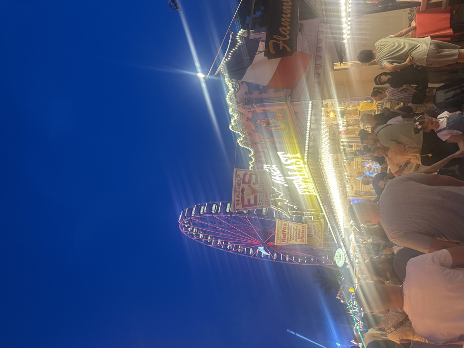 A lively carnival scene unfolds against a dark blue sky. A brightly lit Ferris wheel with red lights spins nearby a busy food stand. The stand has a sign with "Flammkuchen" displayed prominently and a flag. People are milling about, enjoying food and drinks. The crowd appears dense, with many faces and figures partially obscured. The scene is vibrant and full of energy.