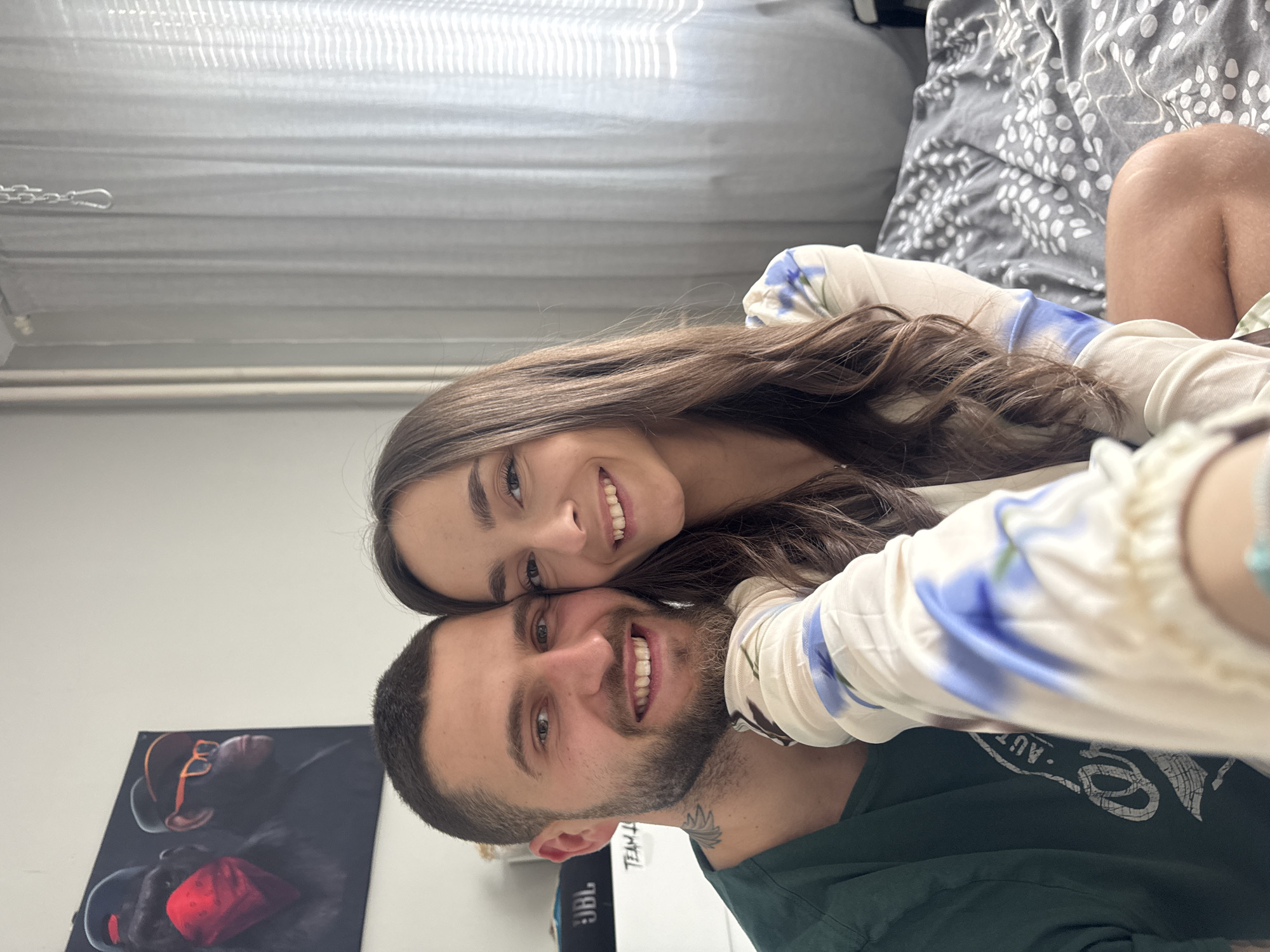 A selfie of a man and a woman. The man has short dark hair and a beard, wearing a green shirt. The woman has long brown hair and is wearing a white top with a floral print. They are smiling and looking at the camera. In the background, there is a wall with a picture of a gorilla wearing glasses, a JBL speaker, a gray curtain, and a bed with patterned sheets. It's not possible to determine the exact location in Sofia, Bulgaria from the visual information provided.