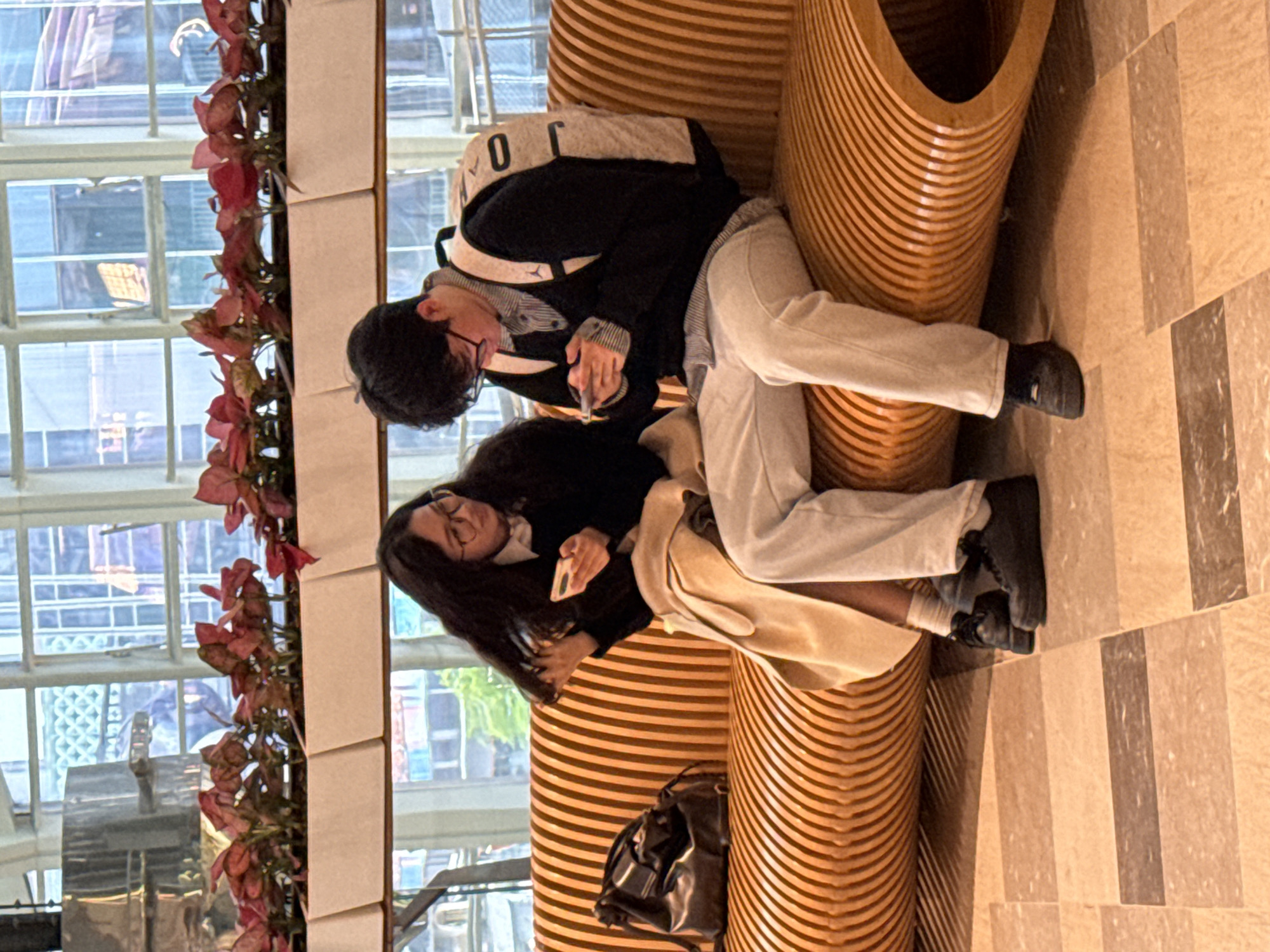 The image depicts an indoor scene in a public space located in Liangjiang, China. Two individuals, one female and one male, are seated side-by-side on a long, curved bench constructed from light brown, vertically ribbed wooden slats.
The female, positioned on the left, has long dark hair and wears eyeglasses. She is dressed in a dark sweater over a white collared shirt, a light beige coat or shawl across her lap, light-colored trousers, and dark footwear. She holds a white smartphone in her left hand and is observed touching her hair with her right hand, her gaze directed towards the phone. A dark-colored shoulder bag rests on the bench to her left.
The male, positioned on the right, has short dark hair and wears eyeglasses. He is dressed in a dark sweater over a striped collared shirt, light-colored trousers, and dark footwear. He wears a light-colored backpack with the number "0" visible on its left side. He holds a dark-colored smartphone with both hands, his head tilted down as he looks at the screen.
Both individuals are independently engaged with their respective mobile devices; no direct interaction between them is observed.
The background features large, multi-paned windows revealing urban buildings in the distance. Below these windows, a row of white rectangular planters contains plants with prominent reddish-pink leaves. The floor is tiled with rectangular sections of varying light brown and beige shades. The scene is brightly lit, primarily by natural light entering through the large windows.
