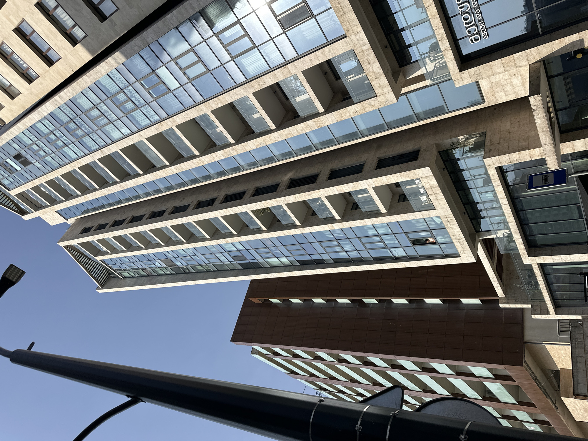 The image is a low-angle shot of modern buildings in Tbilisi, Georgia, captured on a sunny day. One building features a facade of stone and glass panels, with projecting rectangular balconies. Another building has a darker, possibly wood or composite material, exterior. A black streetlight pole is prominently placed in the foreground, obscuring part of the view. The sky is clear and blue. There's a sign visible on one building with Georgian script. A person is visible in the window of the main building, looking down.