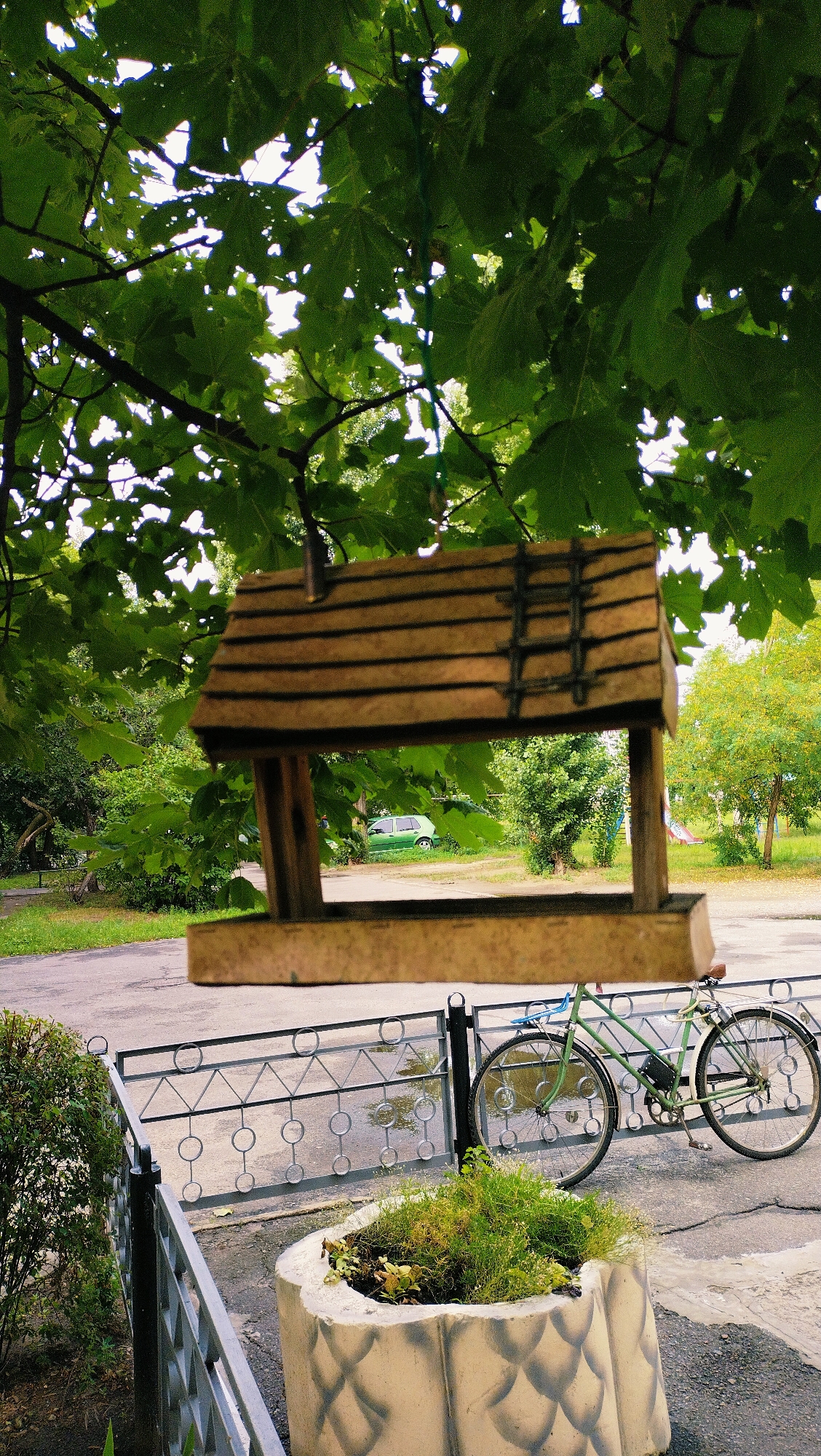 The image depicts an outdoor scene in the city of Horishni Plavni, Ukraine, featuring a wooden bird feeder suspended from tree branches. In the foreground, a concrete planter with greenery sits next to a black metal fence. A light green bicycle leans against the fence, with its front wheel positioned over the fence. Beyond the fence, a paved area is visible, with a green car parked in the distance. The background is filled with trees and foliage, suggesting a park-like or residential area within the city. The overall impression is one of a quiet, somewhat overgrown urban environment.