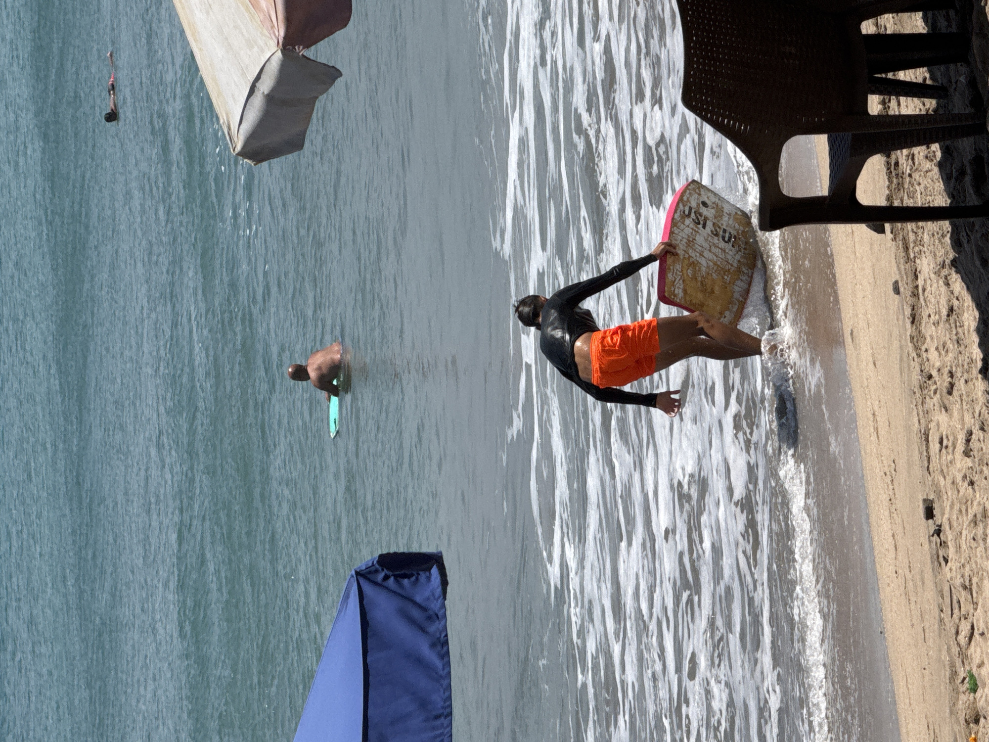 In Kuta, Indonesia, on a beach with tan sand, two individuals are visible in the ocean. In the foreground, a person wearing a black long-sleeved rash guard and bright orange shorts is wading into the foamy surf, carrying a boogie board. The boogie board is pink and yellow and has text printed on it. Behind this person, further out in the turquoise ocean, another individual is visible floating on a green boogie board. In the distance, a third person is seen lying on their back in the water. Two beach umbrellas are also present; a blue one is partially visible on the left and a striped one on the right. A dark brown plastic chair sits on the sand in the foreground. The water is calm with gentle waves.