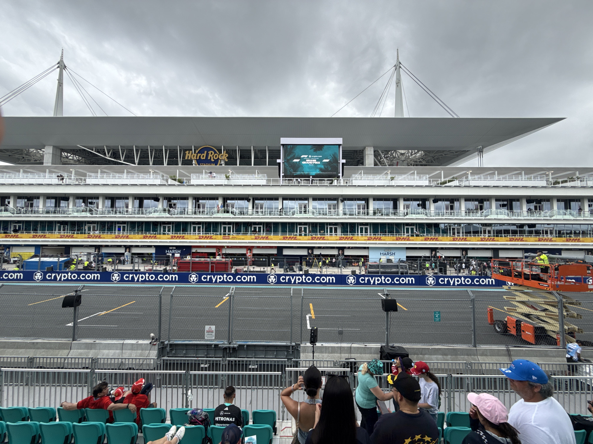 The image shows a view of the Miami International Autodrome, located in Miami Gardens, Florida, United States of America, during the Miami Grand Prix. The track is separated from the seating area by a fence. A large screen displays the event logo and sponsor information. The Hard Rock Stadium is visible in the background. Spectators are seated in the foreground, watching the event. A construction lift with workers is seen on the track level. The environment is overcast with a grey sky.