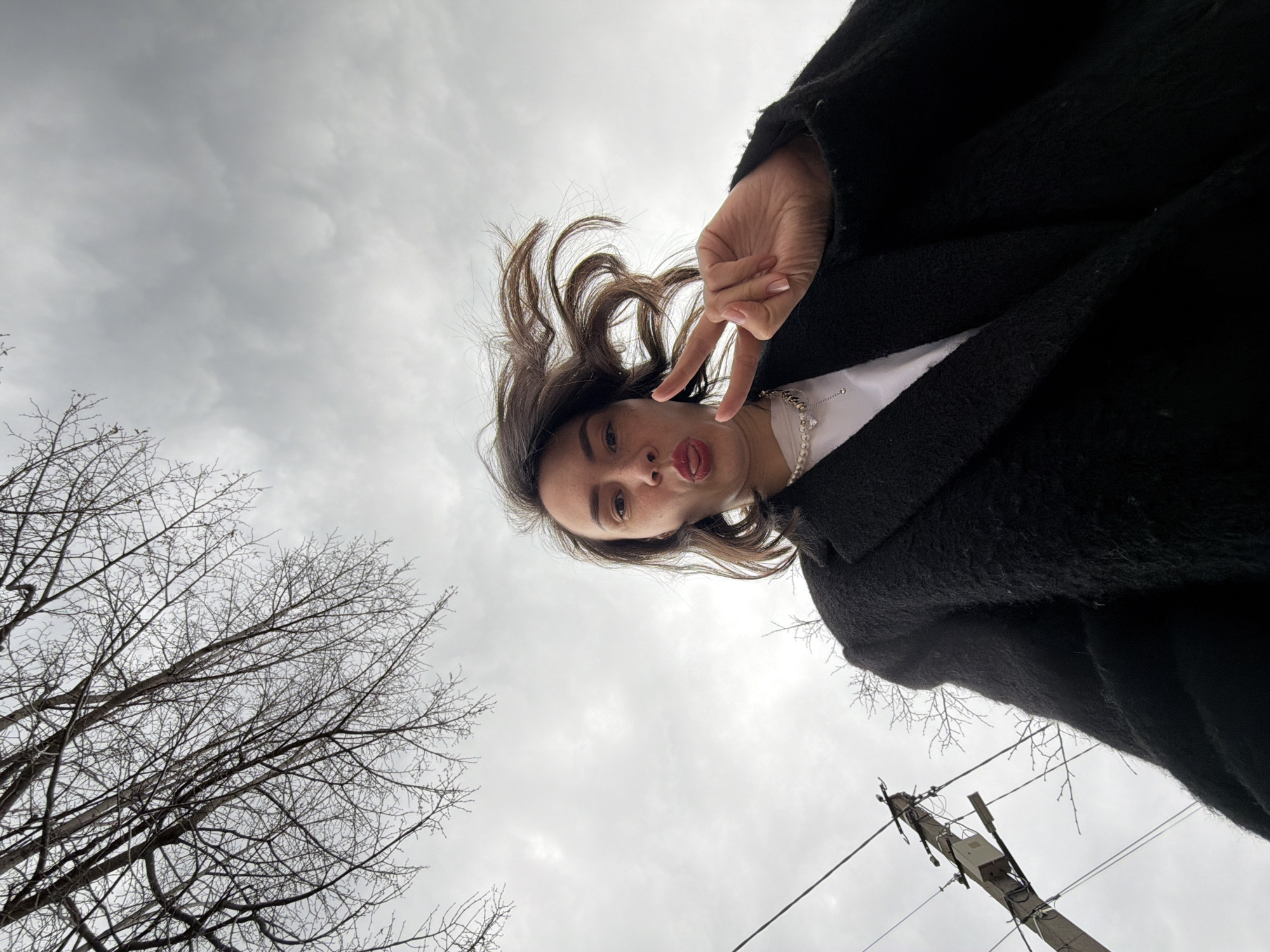 A low-angle shot taken in an outdoor environment, likely in Chaikovsky, Russia. The subject is a young woman with light skin, brown hair, wearing a black coat and a white shirt with a pearl necklace. She is making a peace sign with her right hand, sticking her tongue out. The background features a cloudy sky and bare tree branches on the left, along with a utility pole and wires on the right. The overall setting suggests an urban area during a cold season.