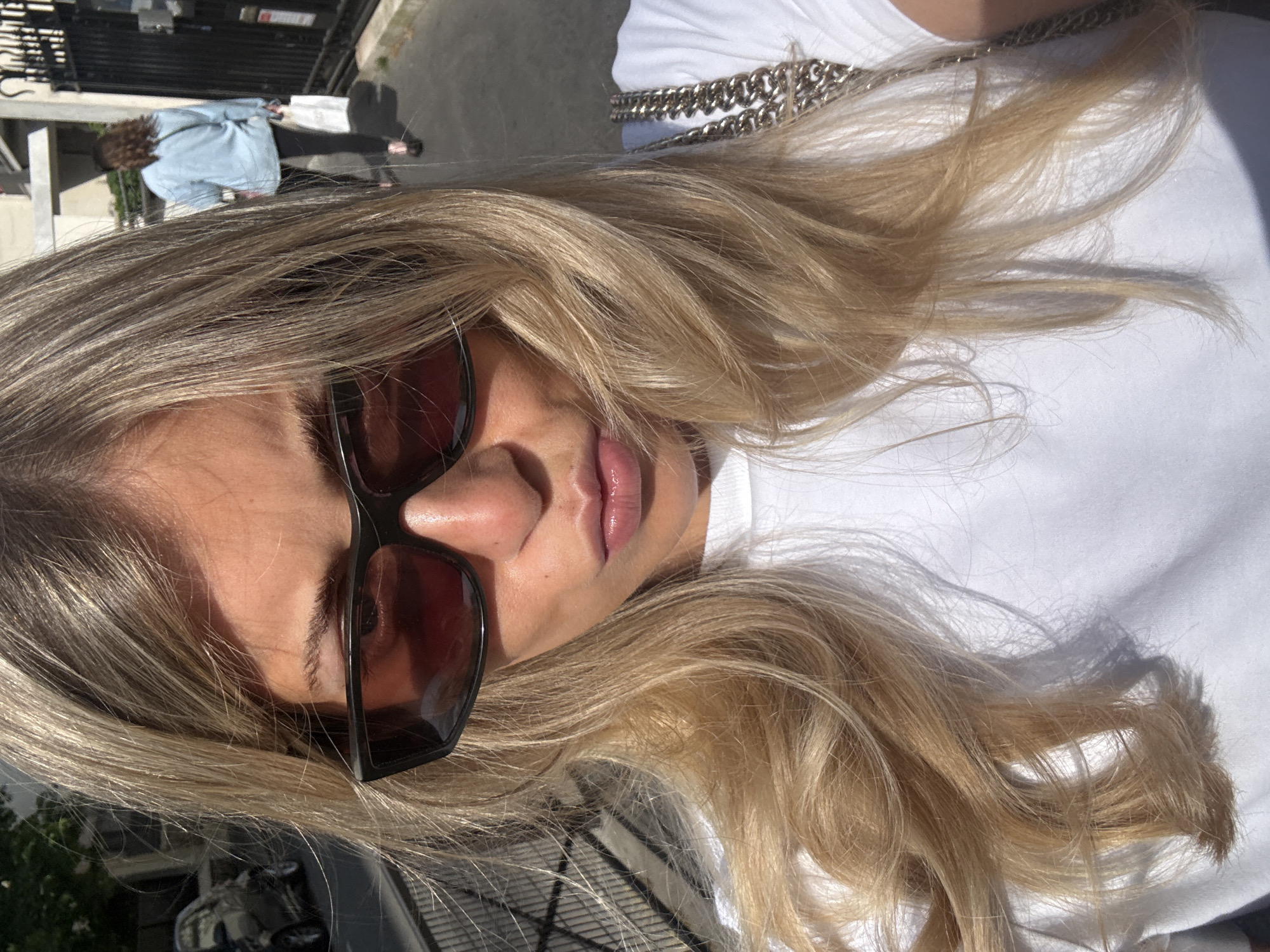 The image is a close-up, tilted portrait featuring a person with long, blonde, wavy hair that cascades around their face and shoulders. The subject is wearing dark-framed sunglasses, which obscure their eyes, and a white t-shirt. A silver chain necklace is visible over the white shirt, partially covered by the hair. Their nose and glosssy, light pink lips are visible, with strong sunlight highlighting the skin.
In the background, out of focus and further away, another person with dark, curly hair is seen from behind, walking. They are wearing a light blue long-sleeved top or jacket and dark trousers. The setting appears to be an urban street or sidewalk, indicated by paved ground, shadows, and a dark, ornate gate or fence in the distance. Reflections of buildings or vehicles can be vaguely seen in the lower left portion of the image.
The lighting suggests a bright, sunny day, with pronounced shadows hinting at a time between late morning and late afternoon. The overall scene conveys a casual, outdoor moment in an urban environment, consistent with a street in Paris, France. There is no visible text in the image.